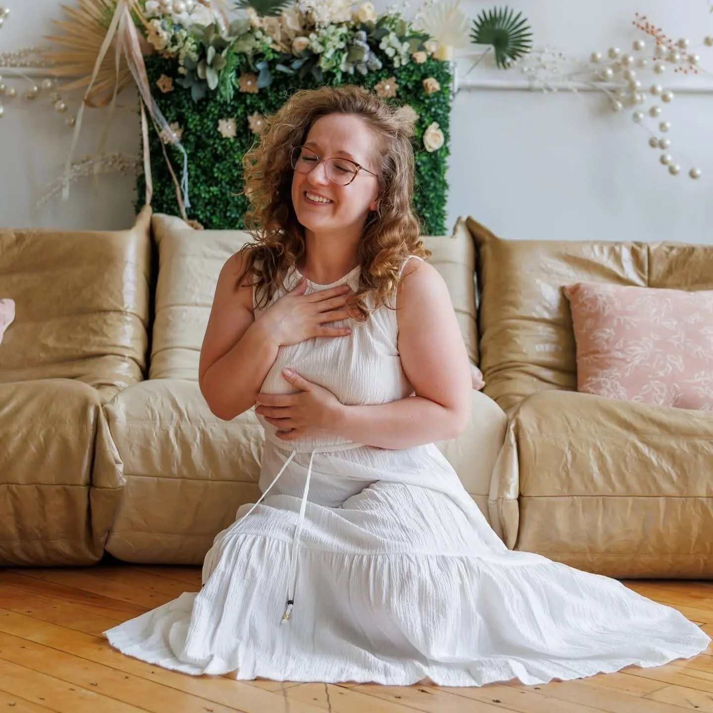 A woman with curly hair and glasses, smiling with eyes closed, sitting on the floor in front of a beige couch, wearing a white dress, with a hand on her chest and another on her stomach, in a decorated room with greenery and floral decorations on the wall behind.