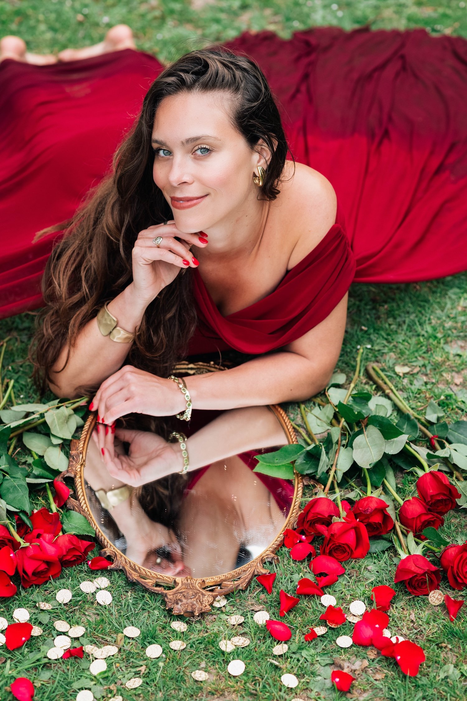 A woman with long dark hair, blue eyes, and wearing gold jewelry is lying on the grass, surrounded by scattered red roses and gold coins. She is looking at the camera with a slight smile and has a mirror reflecting her face on the ground.