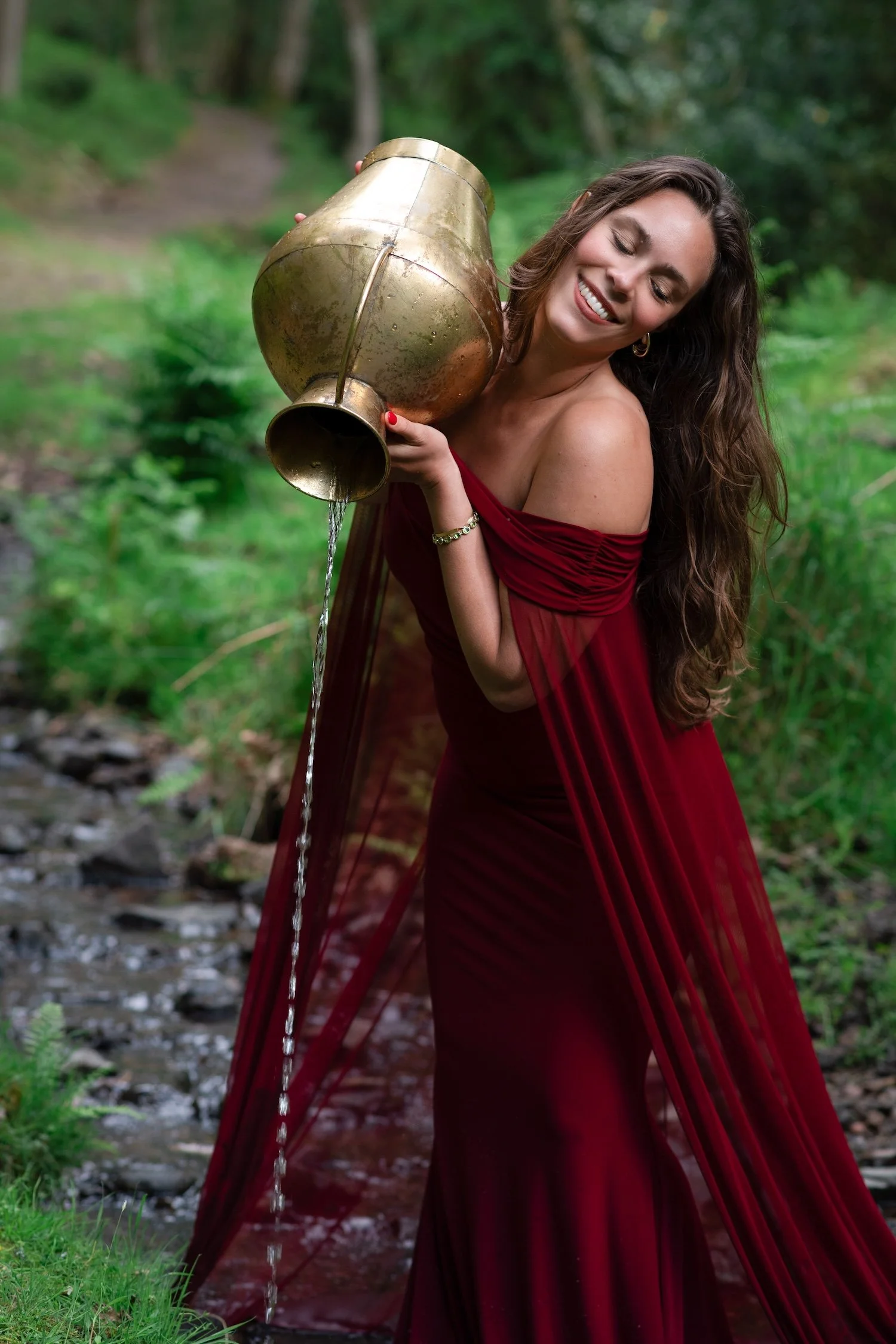 A woman with long dark hair wearing a red dress, holding a vintage brass vase, smiling, in a lush green outdoor setting.