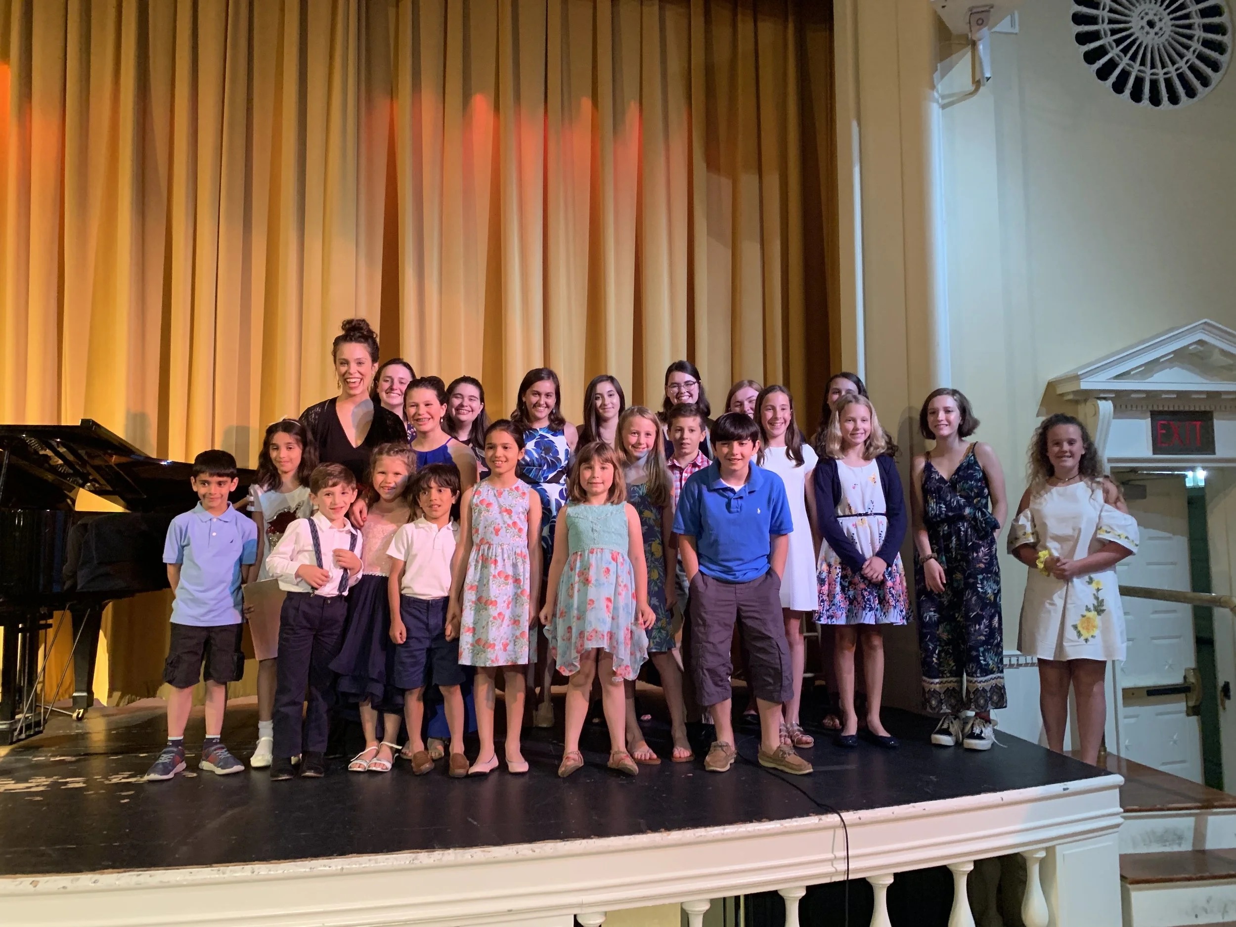 Group of children and adults posing on stage in front of yellow curtains after a performance, with a grand piano visible on the left and an exit sign on the right.
