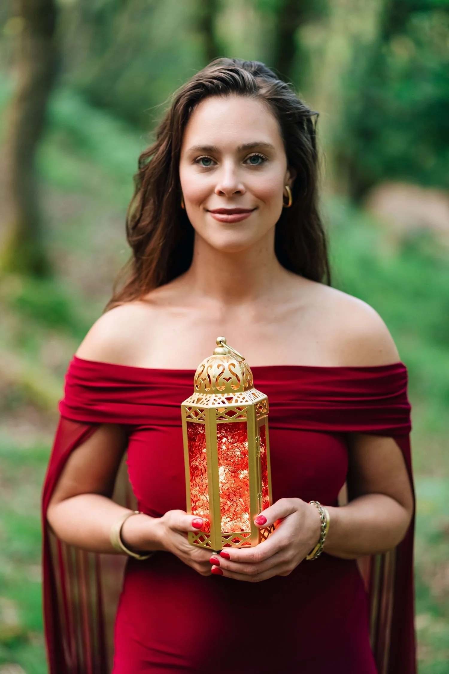 A woman with long brown hair, blue eyes, and hoop earrings wearing a red off-the-shoulder dress, standing outdoors in a lush green forest, holding a lit decorative lantern with a golden structure and red glass panels.