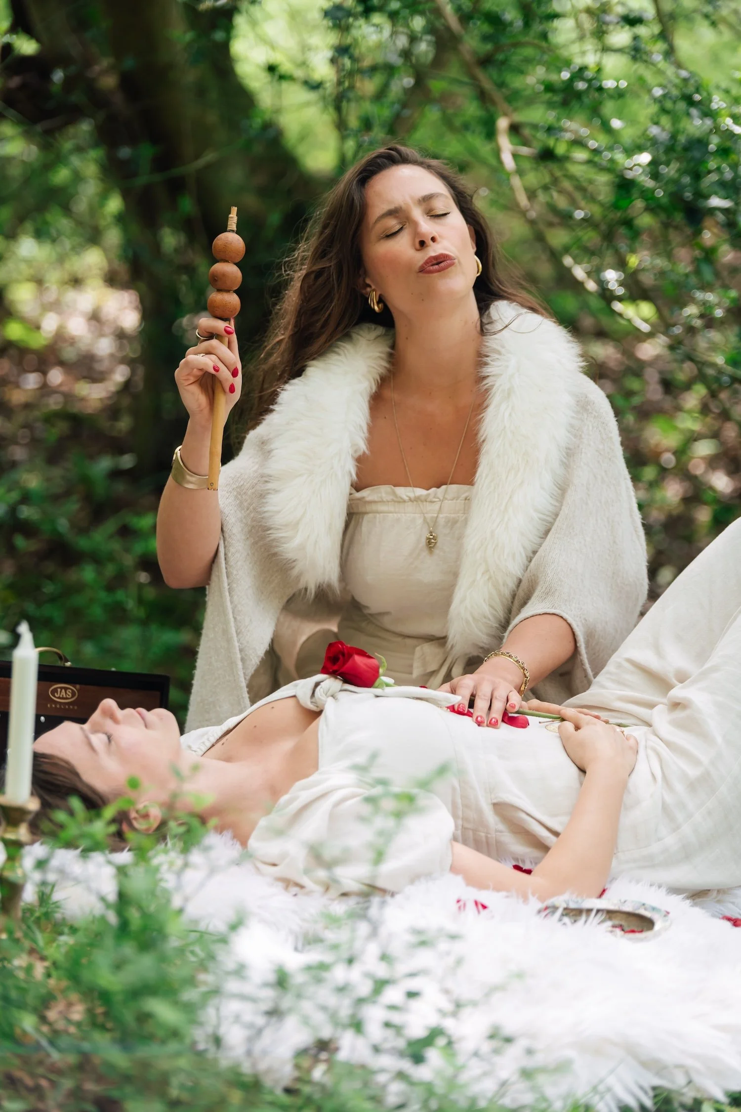 A woman in a white dress lying on a fluffy white surface with a red rose. A woman standing nearby is holding a skewer with four round foods, possibly olives or pickles, and is wearing a white fur-lined jacket.