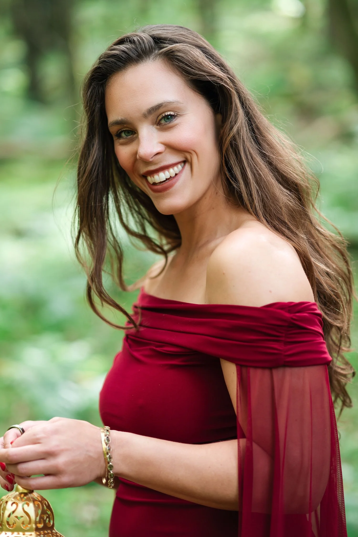 A woman with long brown hair, blue eyes, and light skin smiling outdoors in a green, leafy setting. She is wearing a burgundy off-the-shoulder dress with sheer sleeves and jewelry on her wrist, holding a decorative gold Christmas ornament.