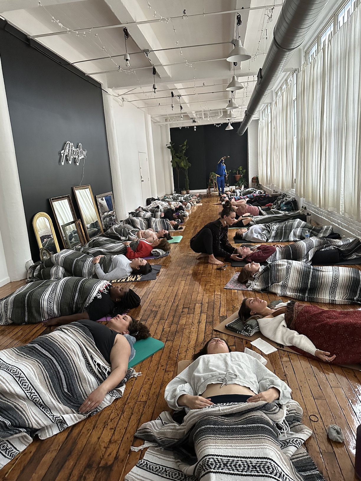 A group of people participating in a yoga or meditation class in a spacious room with wooden floors and large window drapes. Participants are lying down, covered with blankets, and some are being guided by an instructor.