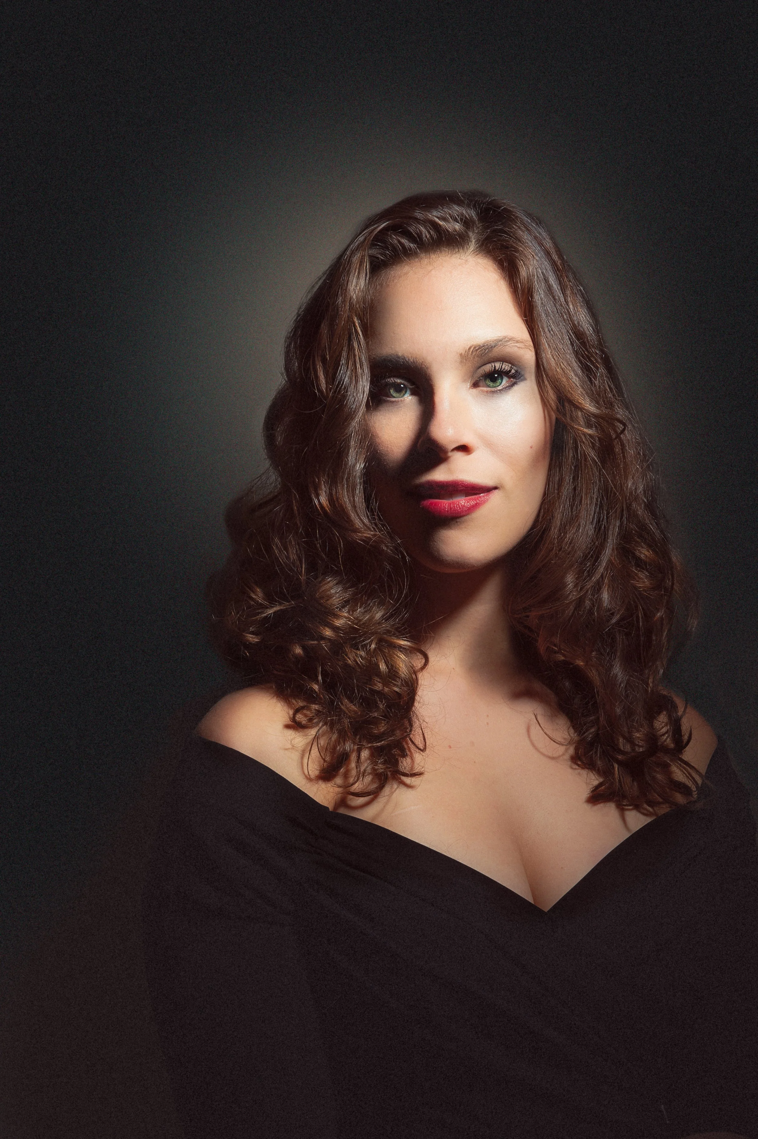 A woman with long, curly brown hair and fair skin, wearing a black off-the-shoulder top, looking directly at the camera against a dark background.