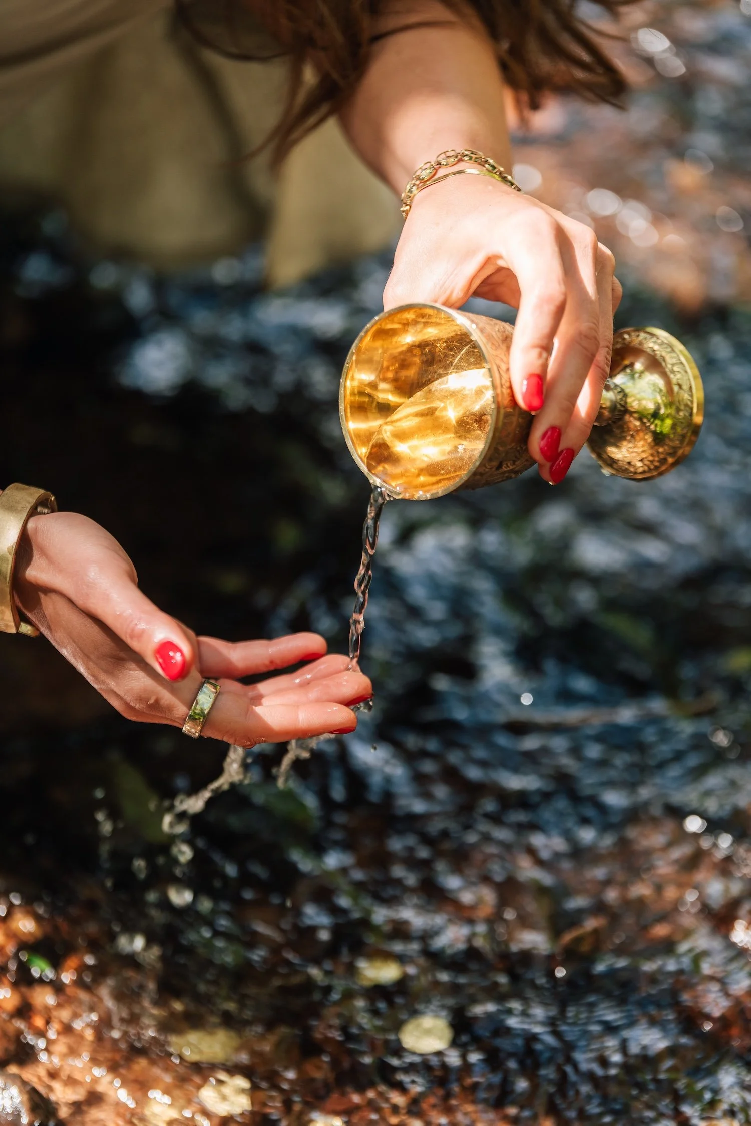 A person with red hair pouring water from a golden cup into their hand, outdoors near a stream or river, with the water flowing over rocks.