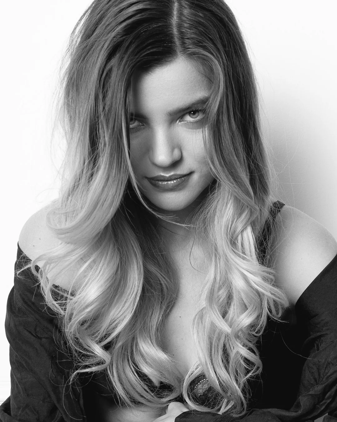 Portrait en noir et blanc d’une femme au regard perçant, avec de longs cheveux ondulés et un léger sourire. Son épaule dénudée et sa tenue suggèrent une allure à la fois sensuelle et mystérieuse.