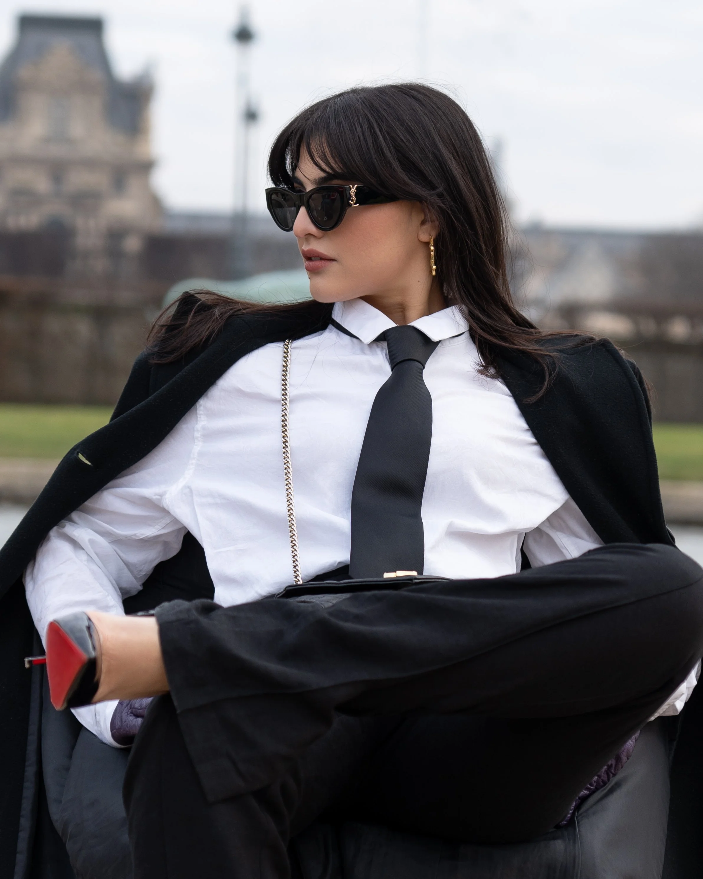Femme élégante en chemise blanche, cravate noire, pantalon et manteau noirs, portant des lunettes de soleil noires. Assise, jambe croisée, regardant sur le côté, elle pose à l'extérieur avec un bâtiment en arrière-plan.