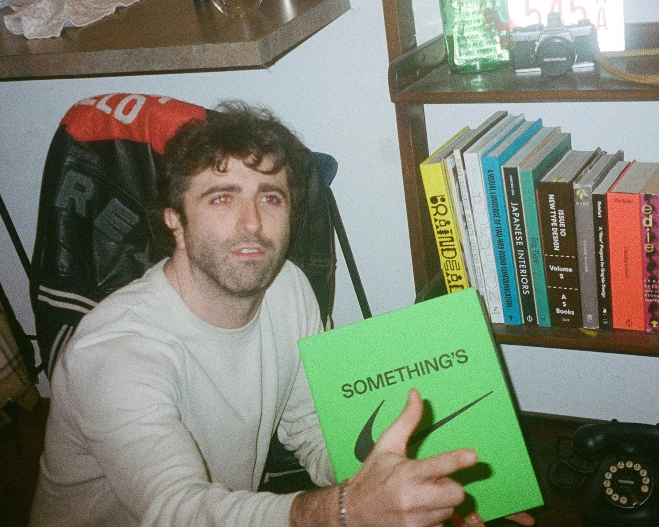 A man with dark curly hair and a beard sitting in a black chair with red and black clothing draped over the back. He is holding a bright green book titled 'SOMETHING'S' with a stylized logo, and is looking slightly upwards. Behind him, there is a bookshelf with various books and a dark-colored desk with a vintage rotary phone and a black vintage camera.