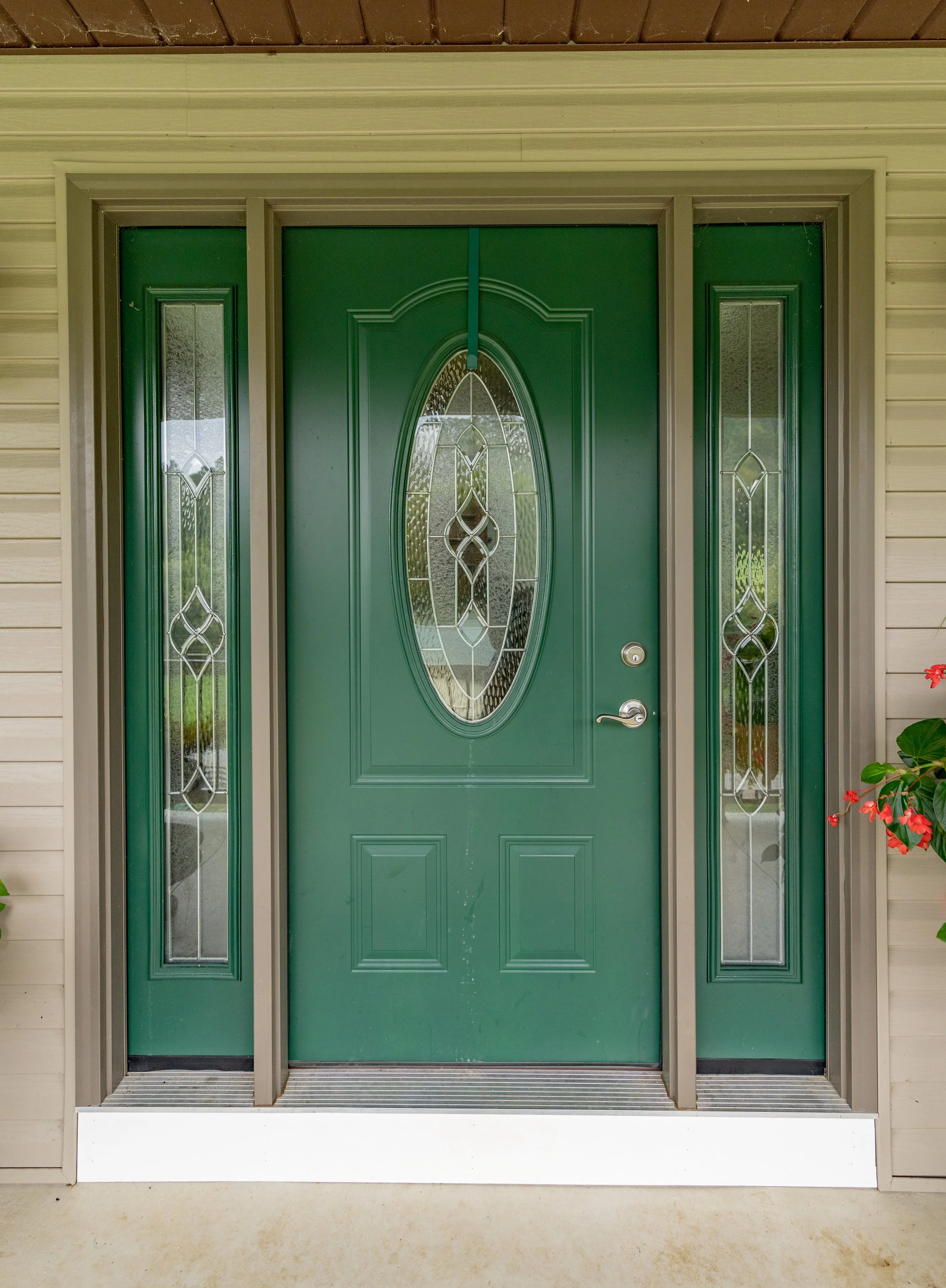 Front door of a house with double wooden doors, brass handles, flanked by paneled windows, and flanked by black shutters. The exterior features grey siding, wall-mounted lanterns, a mail slot, and a small garden with plants.