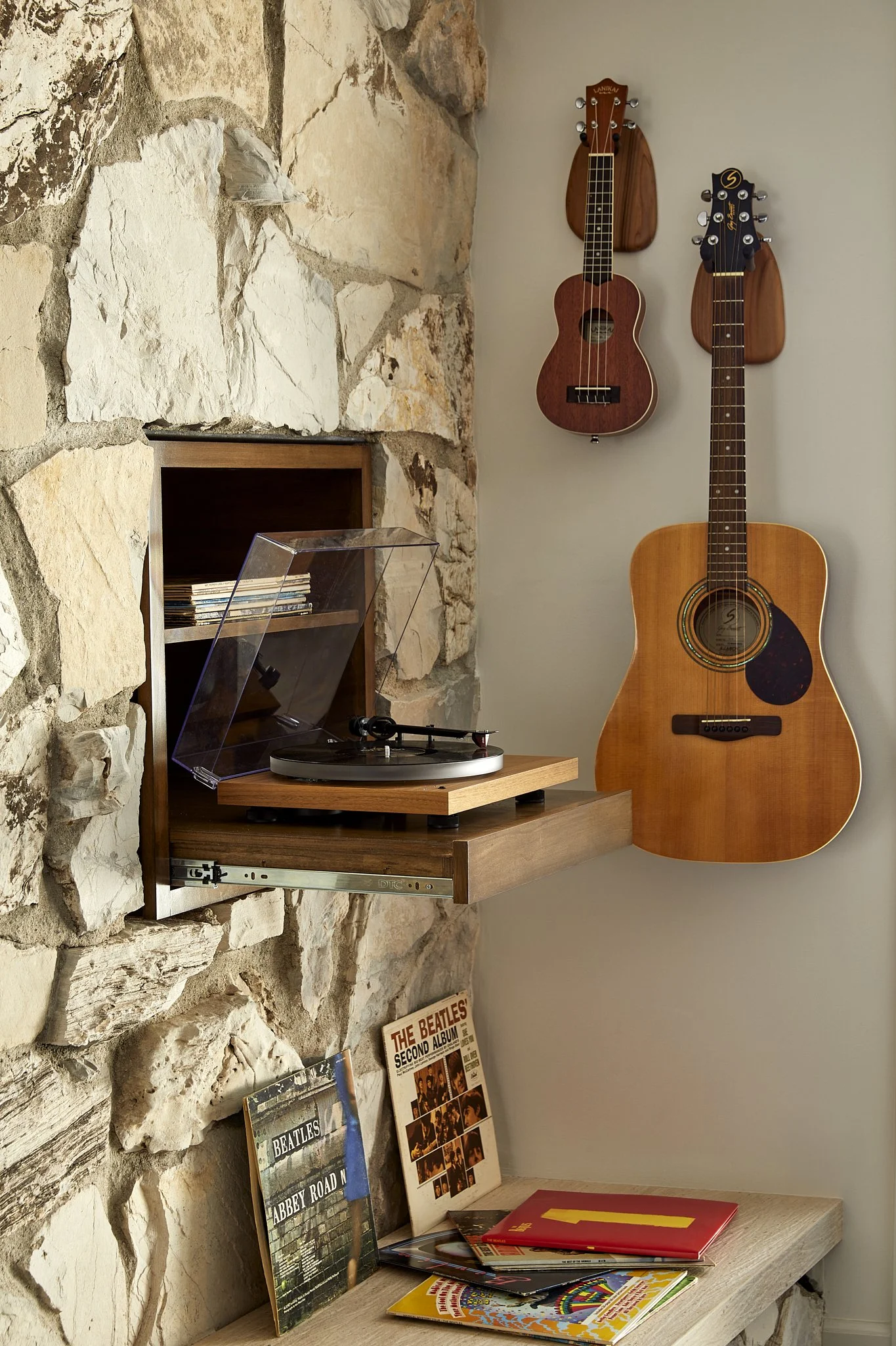A turntable with a clear plastic cover on a wooden shelf next to a stone fireplace. Two acoustic guitars hang on the wall above, and a pile of vinyl records rest on a wooden surface below.