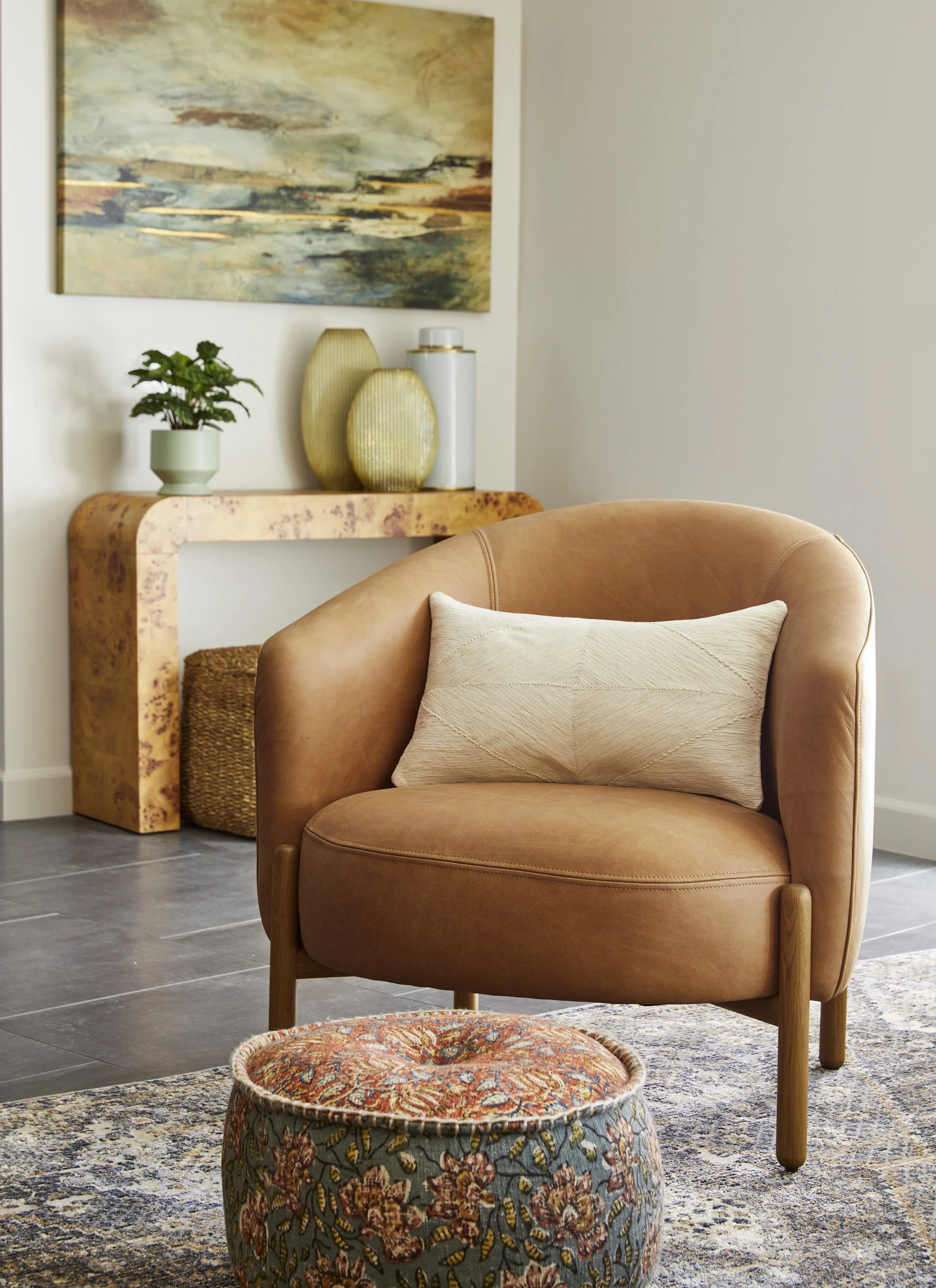 A cozy living room corner with a tan leather armchair, a beige pillow, a patterned round ottoman, a wooden console table, a potted plant, decorative vases, and an abstract painting on a white wall.
