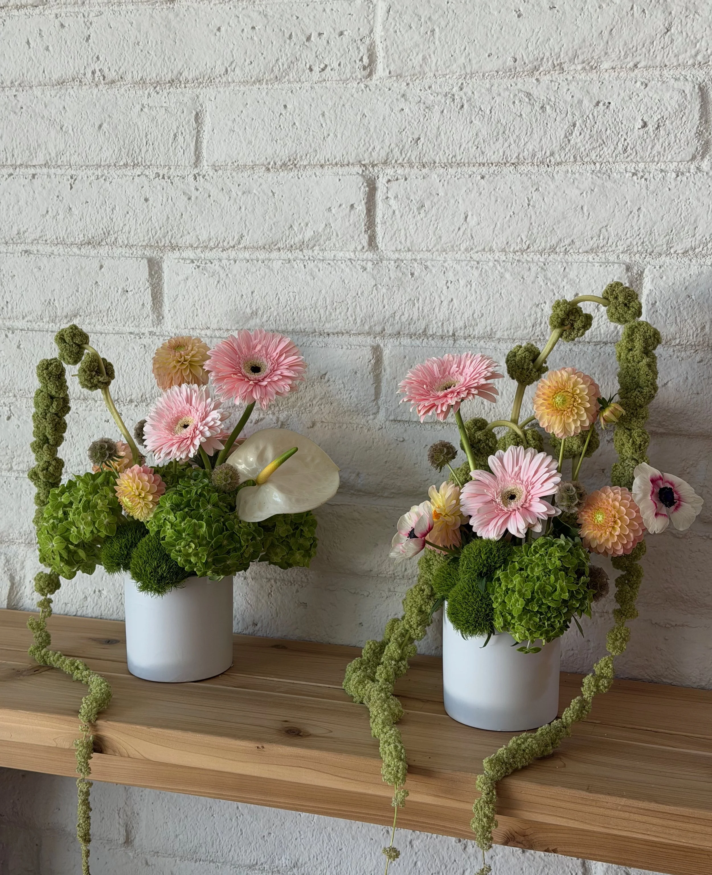 Two floral arrangements with pink gerbera daisies, anthurium, green hydrangea, and trailing amaranthus in white ceramic vessels from a Bloom Bar LA flower arranging workshop in Los Angeles.