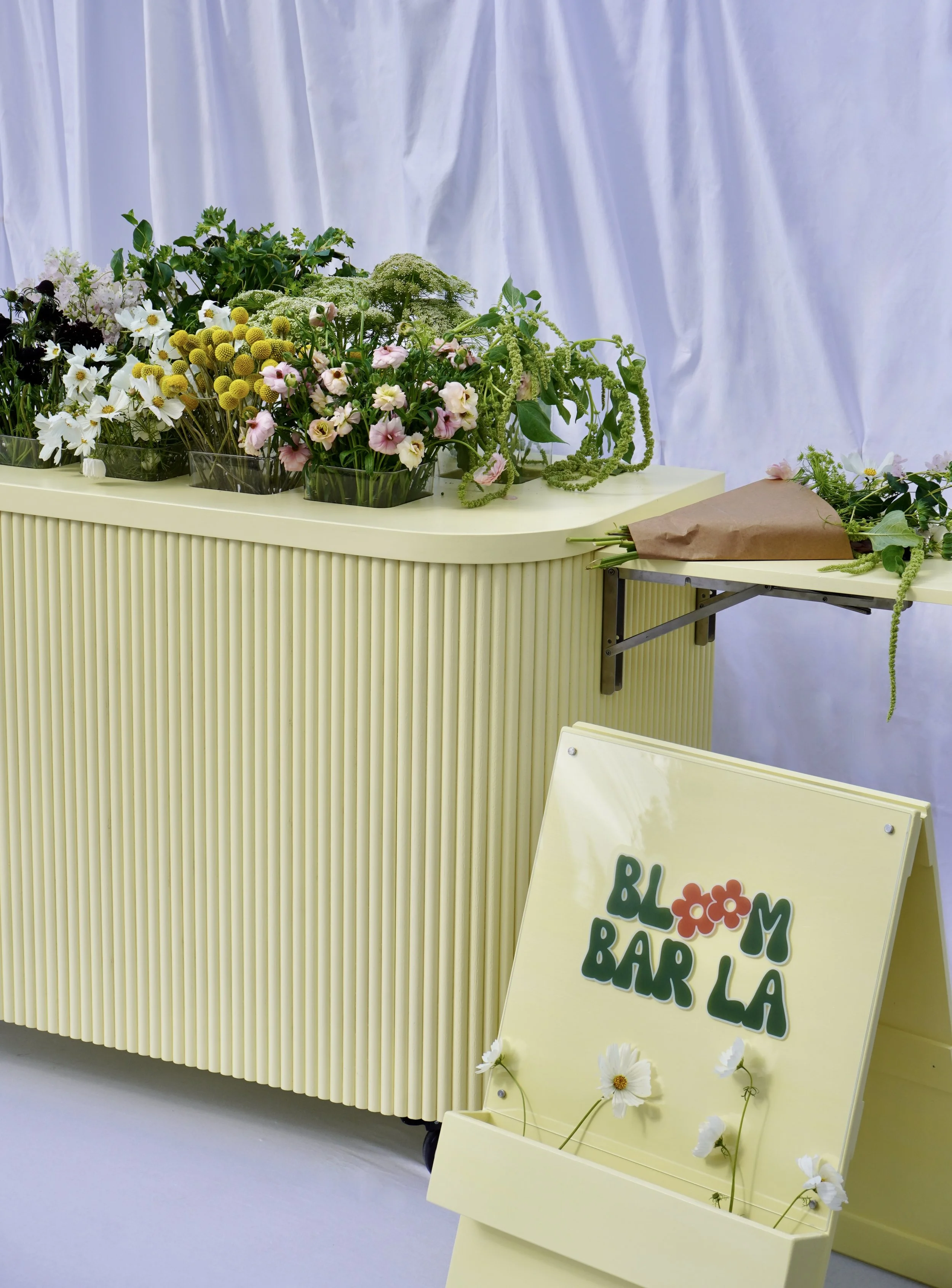 Bloom Bar LA pop-up flower bar fully set up with an abundant selection of seasonal stems and a side wrapping station, displayed against a white draped backdrop with branded A-frame signage.