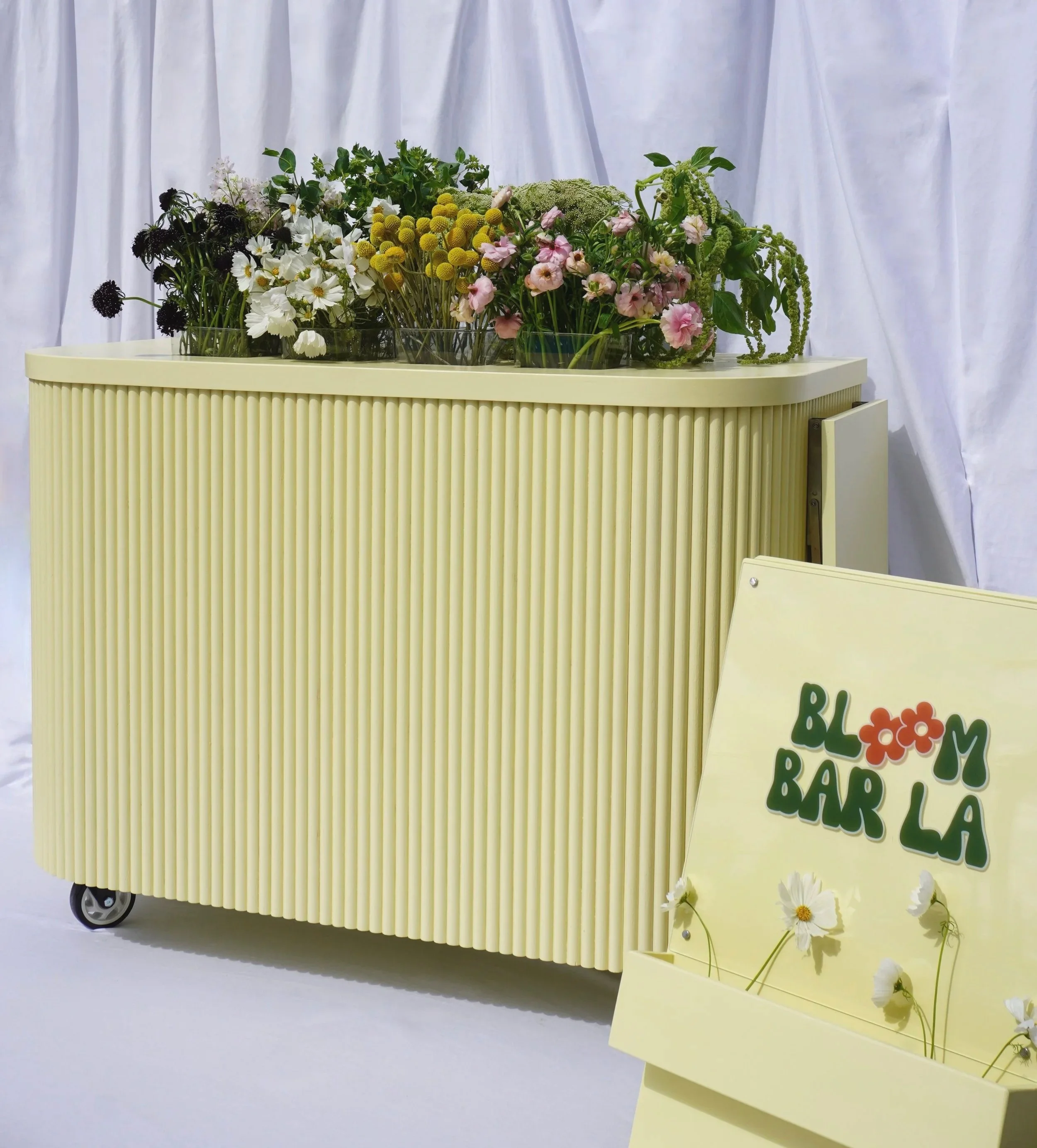 loom Bar LA mobile flower bar cart styled with fresh seasonal blooms including cosmos, ranunculus, and craspedia against a white draped backdrop, with branded signage in the foreground.