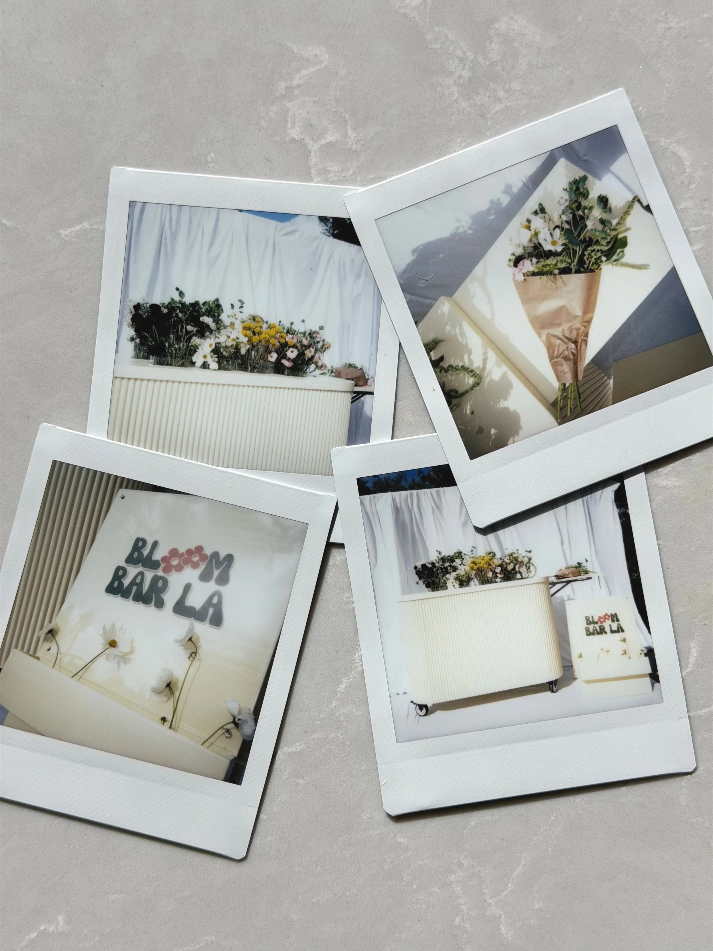 A flat lay of four Polaroid photos showcasing the Bloom Bar LA pop-up flower bar setup including fresh bouquets, branded signage, and floral arrangements at a Los Angeles event.