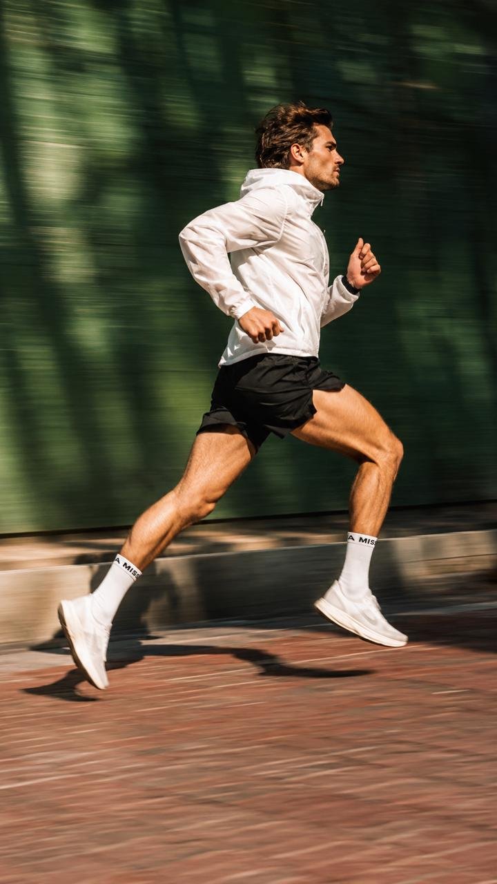 A man wearing a white jacket, black shorts, and white running shoes is running fast outdoors on a brick path, with a blurred green background indicating motion.