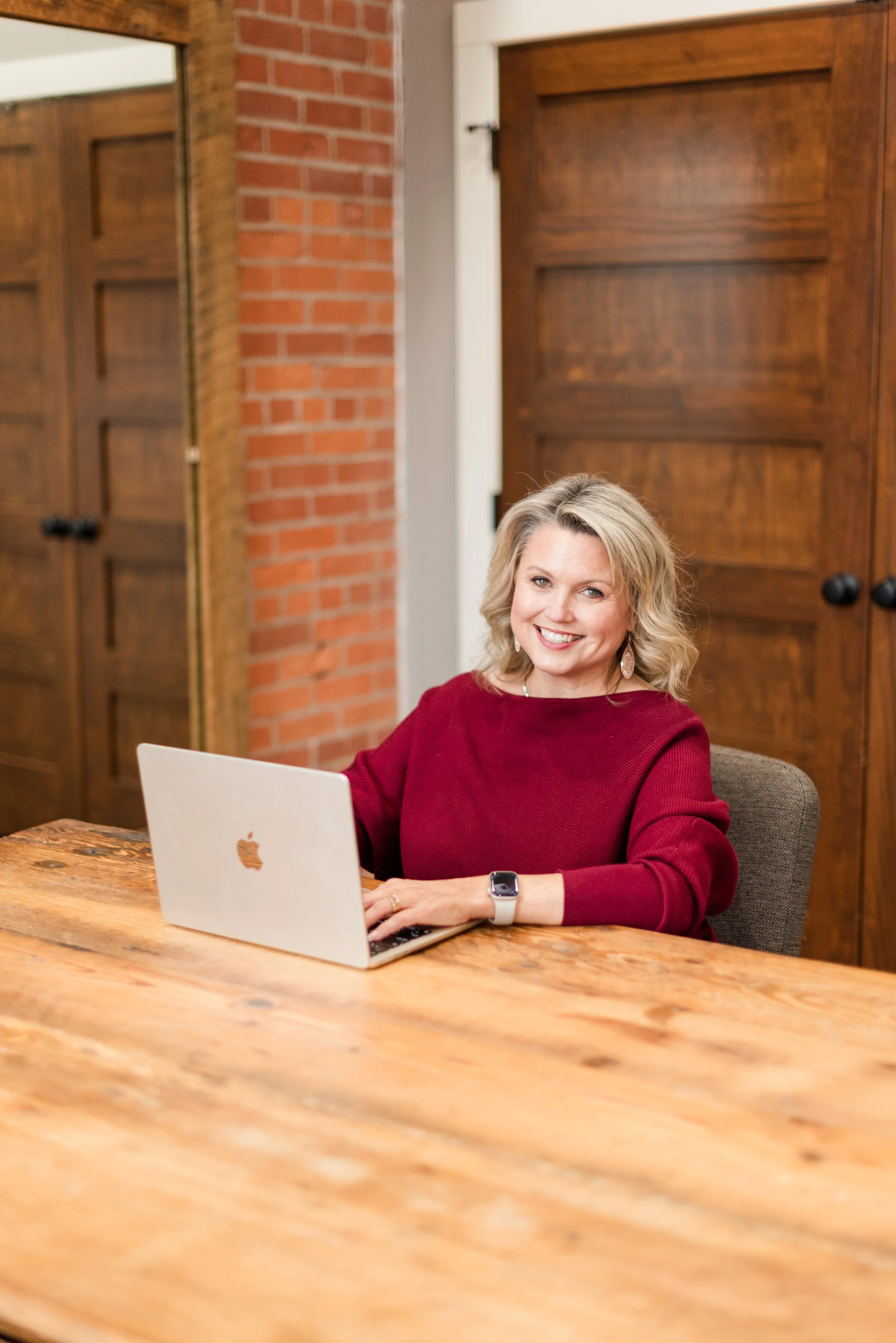 Woman in a red sweater using a laptop at a wooden table in an office setting.