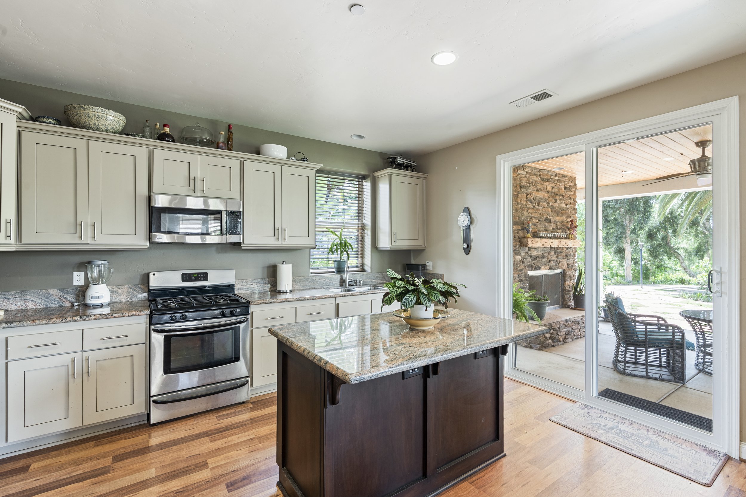Kitchen with beige cabinets, marble countertops, and a small island. A sliding glass door leads to a patio with outdoor seating and a stone fireplace.
