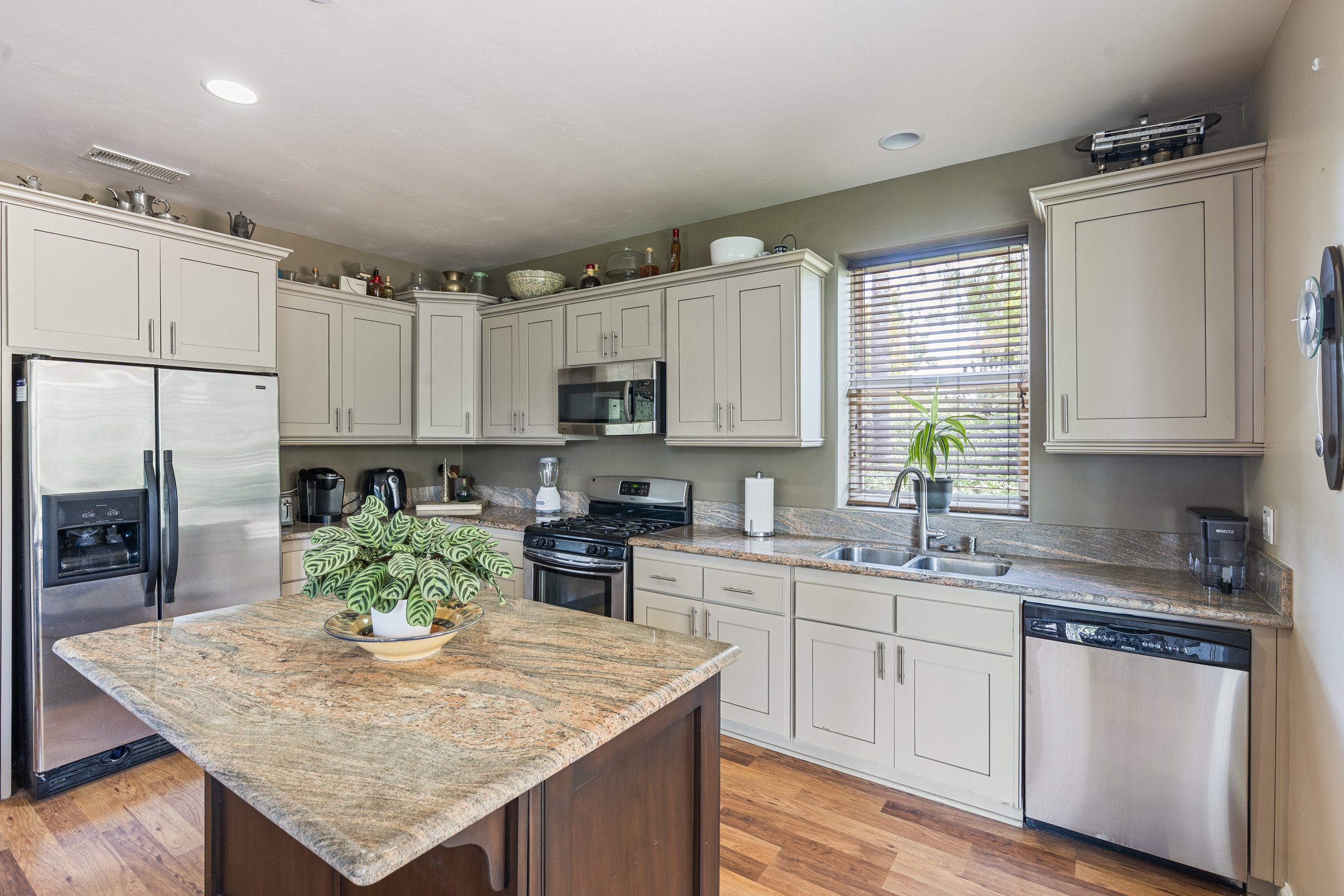 Kitchen with white cabinets, granite countertops, stainless steel appliances, a window with blinds, and a small wooden island with a potted plant.