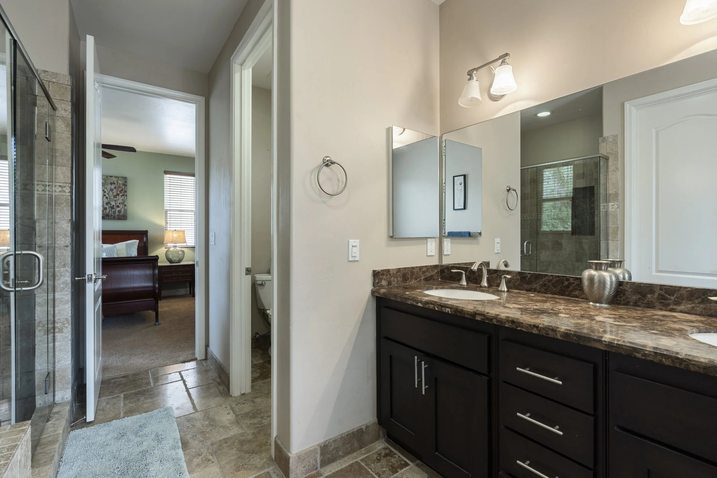 Bathroom with dark wood vanity, marble countertop, large mirror, two sinks, silver fixtures, and decorative vases; glass shower with beige tile, adjacent to a toilet and bedroom with bed and nightstand.