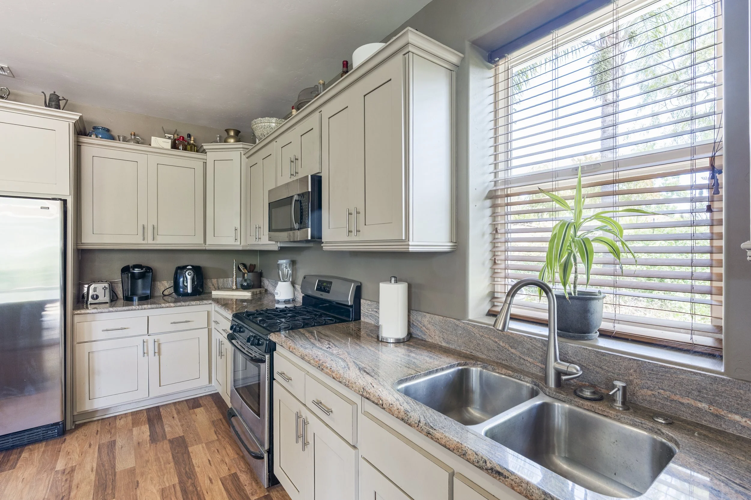 Kitchen with white cabinets, granite countertops, a stainless steel stove, microwave, dishwasher, and a large window with wooden blinds and a potted plant.