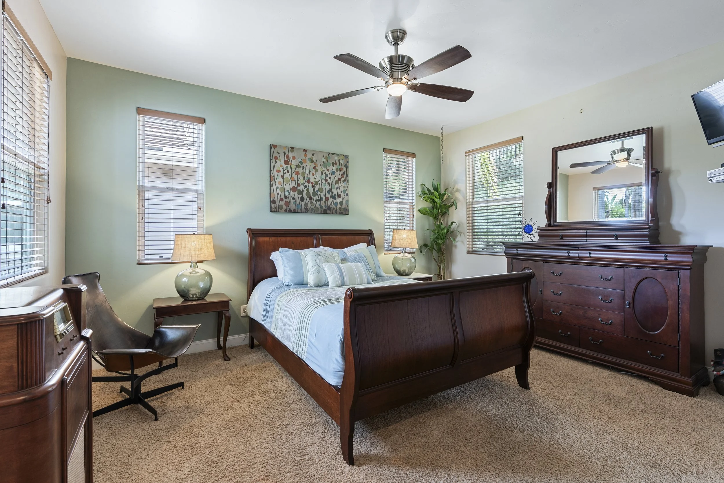 Bedroom with wooden bed, dresser with mirror, leather chair, nightstand, lamps, painting, ceiling fan, and multiple windows with blinds.