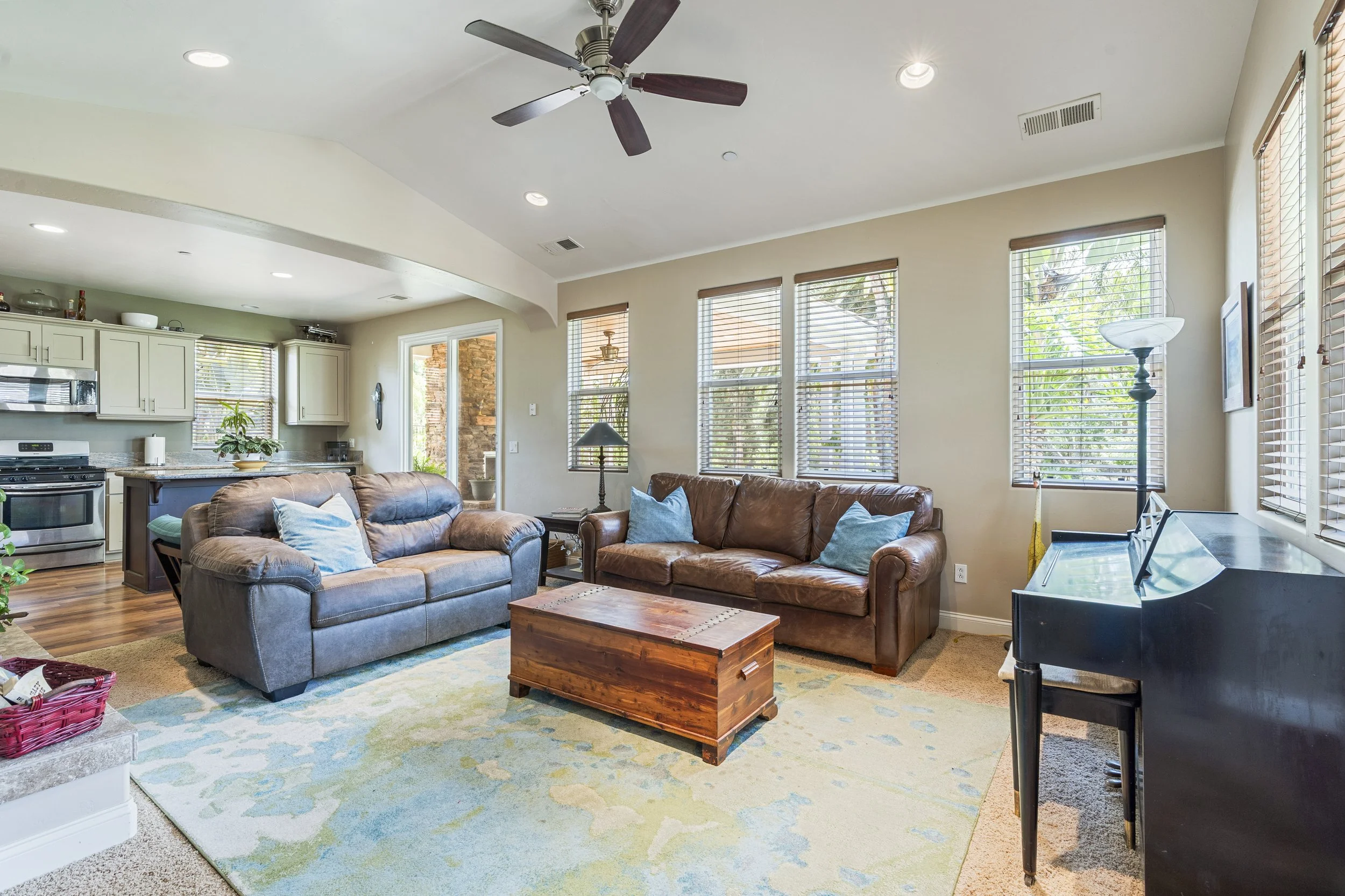 Living room with two brown leather sofas, a wooden coffee table, a black piano with a bench, a floor lamp, and multiple windows with blinds, adjacent to an open kitchen with white cabinets and a small island.