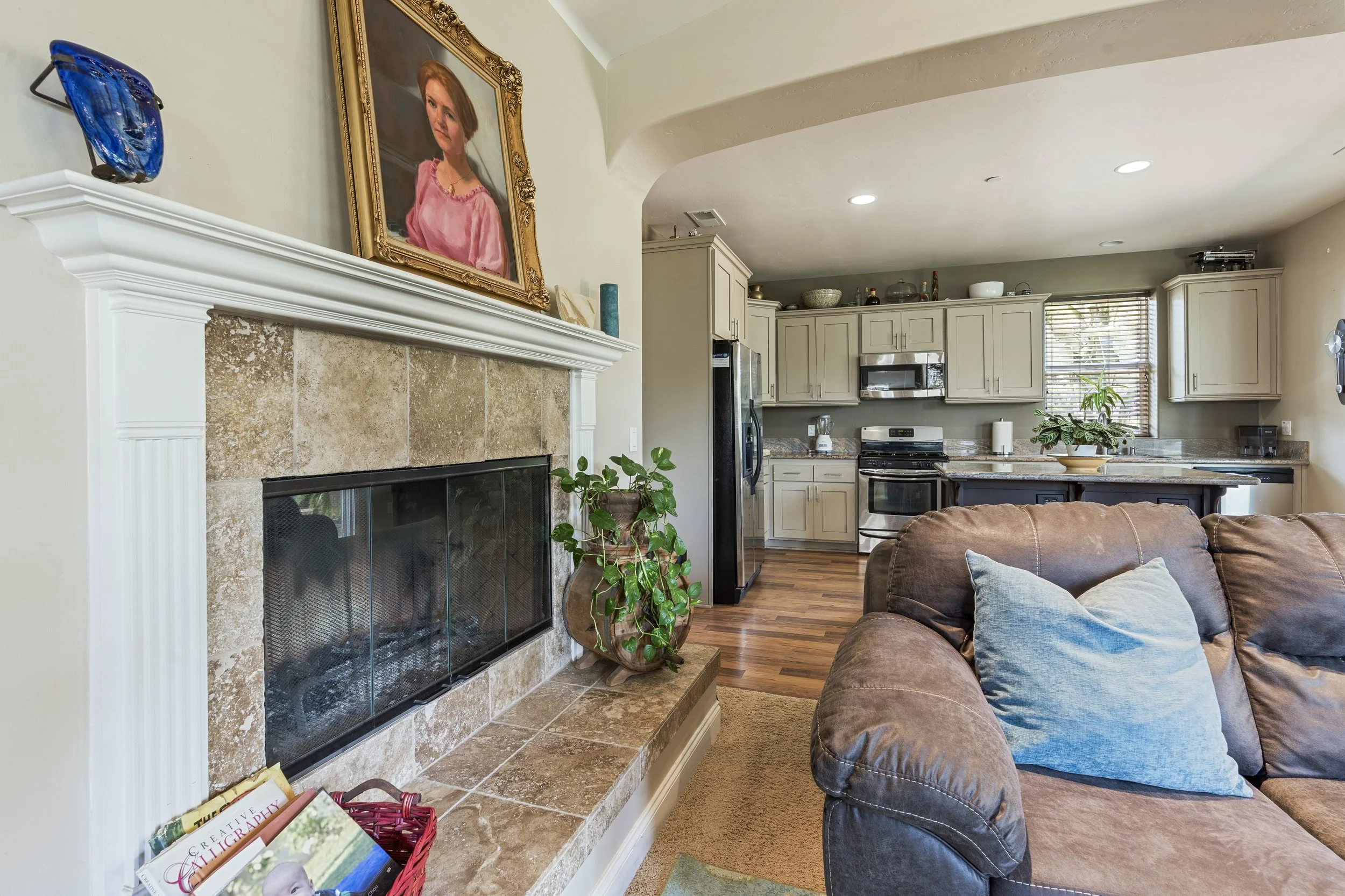 Living room with a fireplace, framed portrait above, couch with a blue pillow, and open kitchen in the background with gray cabinets and potted plants.