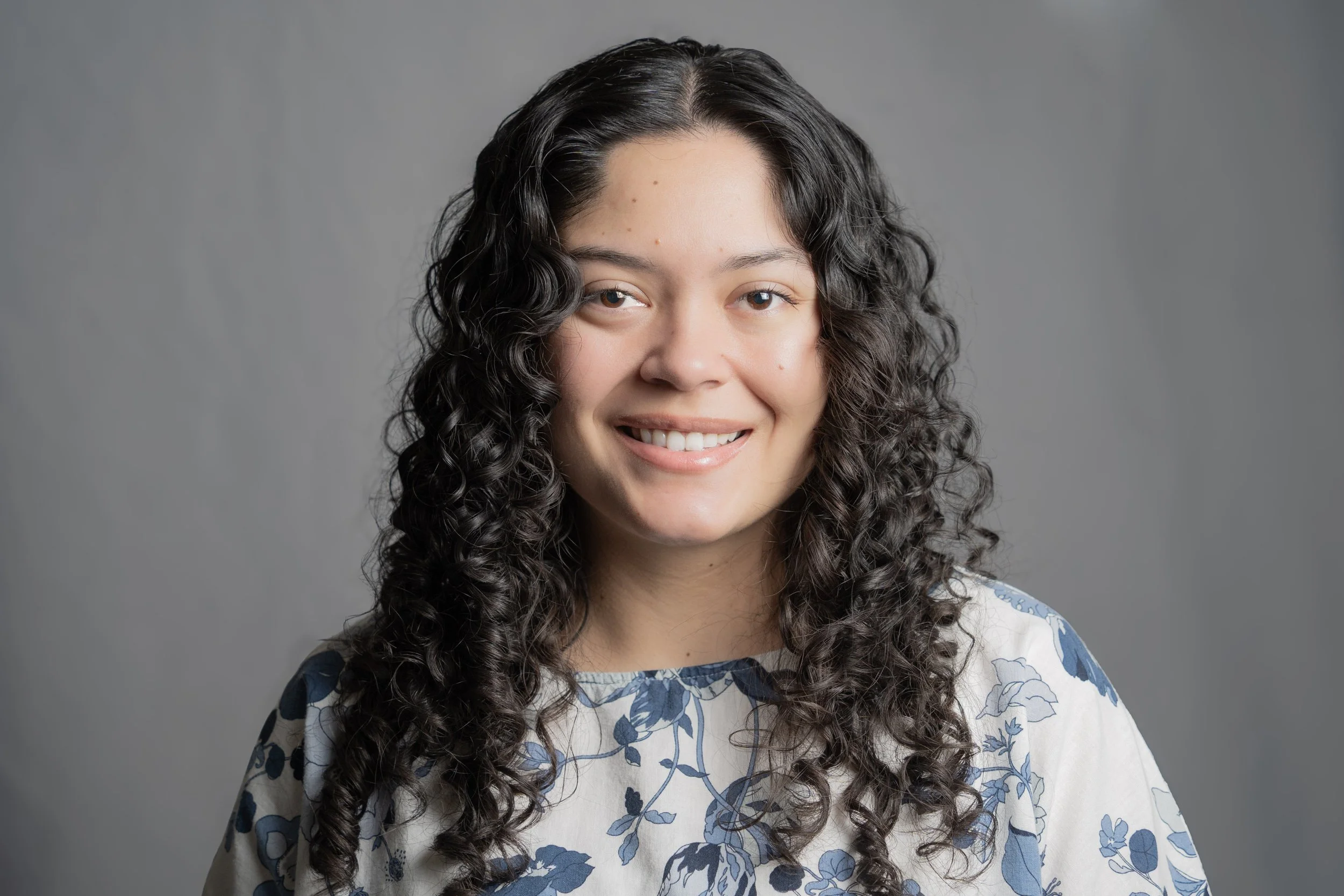 A young woman with curly brown hair, blue eyes, and a light complexion smiling at the camera. She is wearing a white top with blue floral patterns and is set against a gray background.