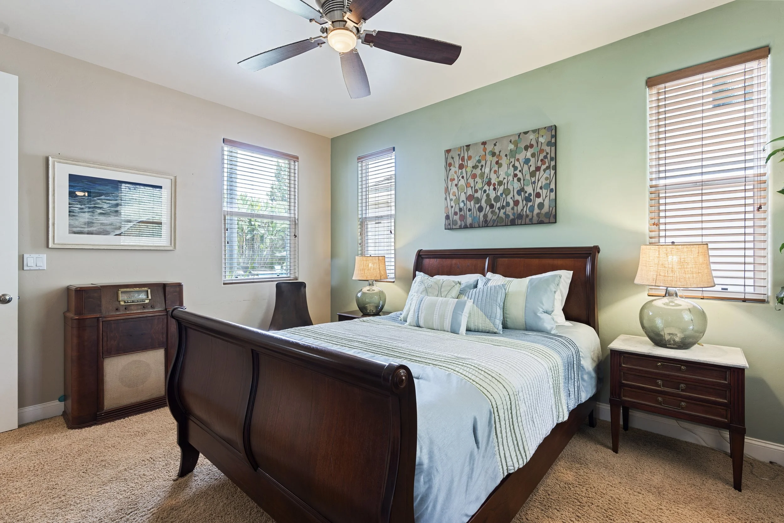 Bedroom with green and beige walls, wooden bed with beddings, nightstands with lamps, ceiling fan, three windows with blinds, and a painting of colorful abstract flowers above the bed.