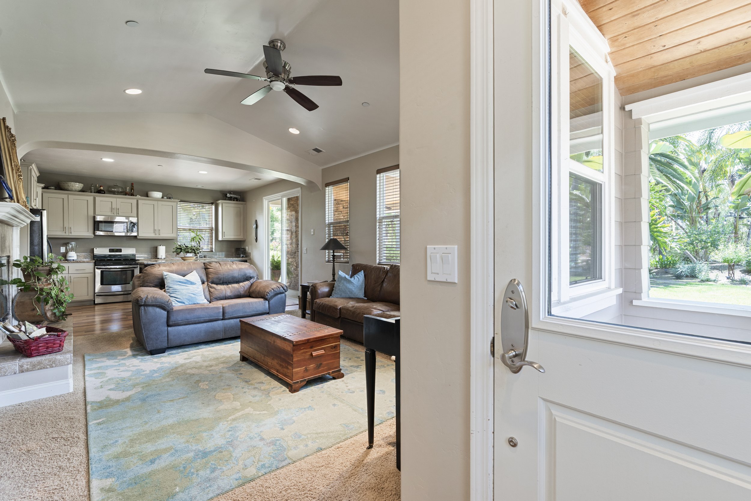 Living room with beige walls and ceiling, featuring two sofas, a wooden coffee table, a fireplace, and large windows showing greenery outside, seen from the front door.