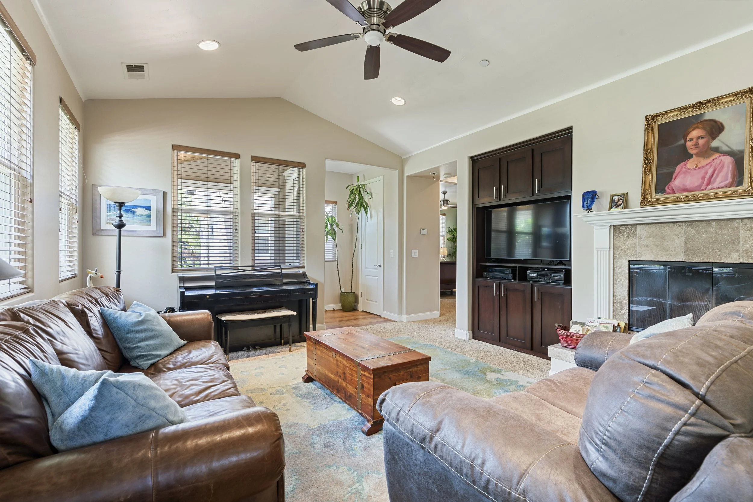 Living room with brown and beige couches, a wooden coffee table, a black piano, a large flat-screen TV, and a fireplace with a portrait above it, illuminated by multiple windows with blinds.