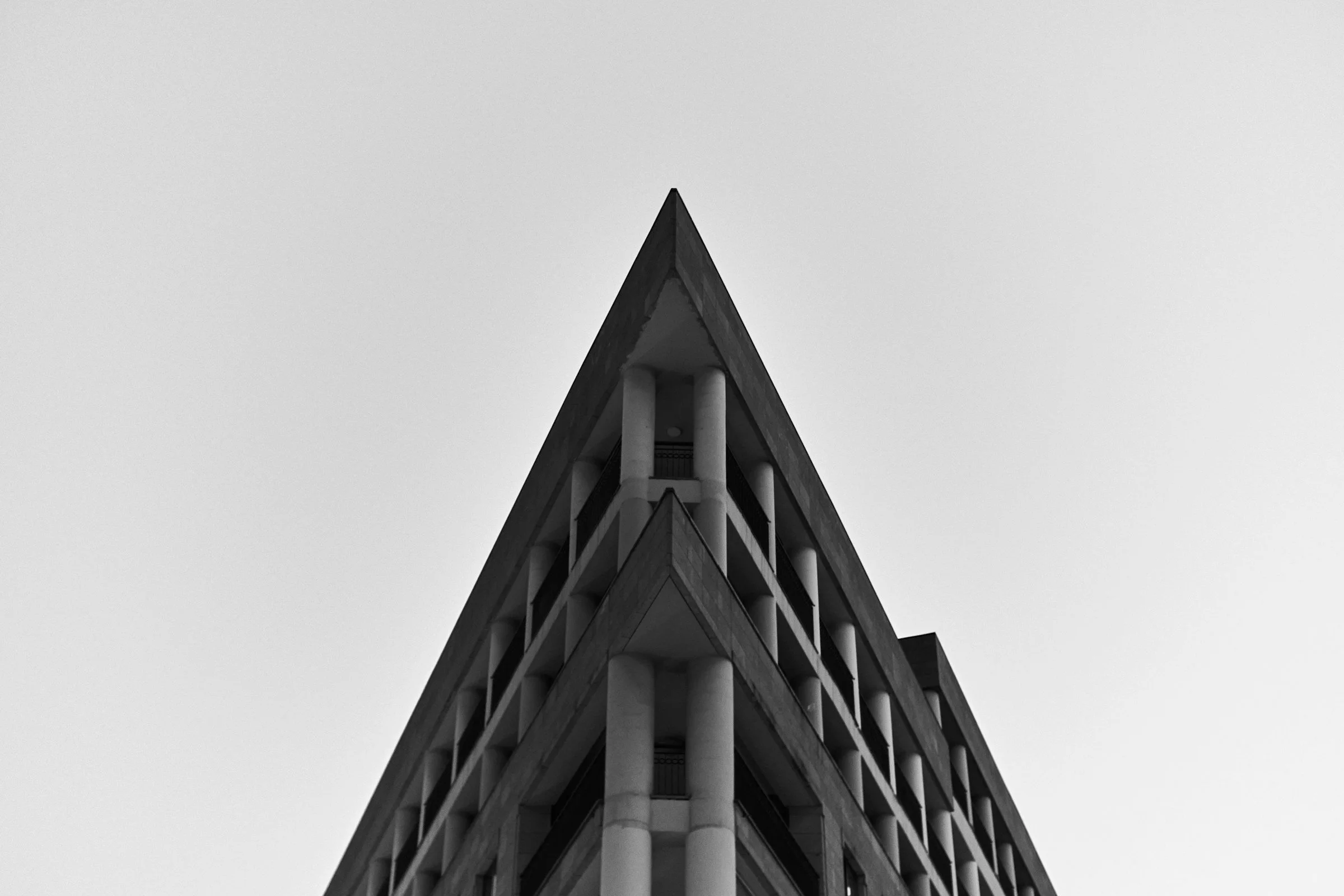 Black and white photograph of the corner of a modern multi-story building with columns, sharp angles, and grid-like windows, against a blank sky.