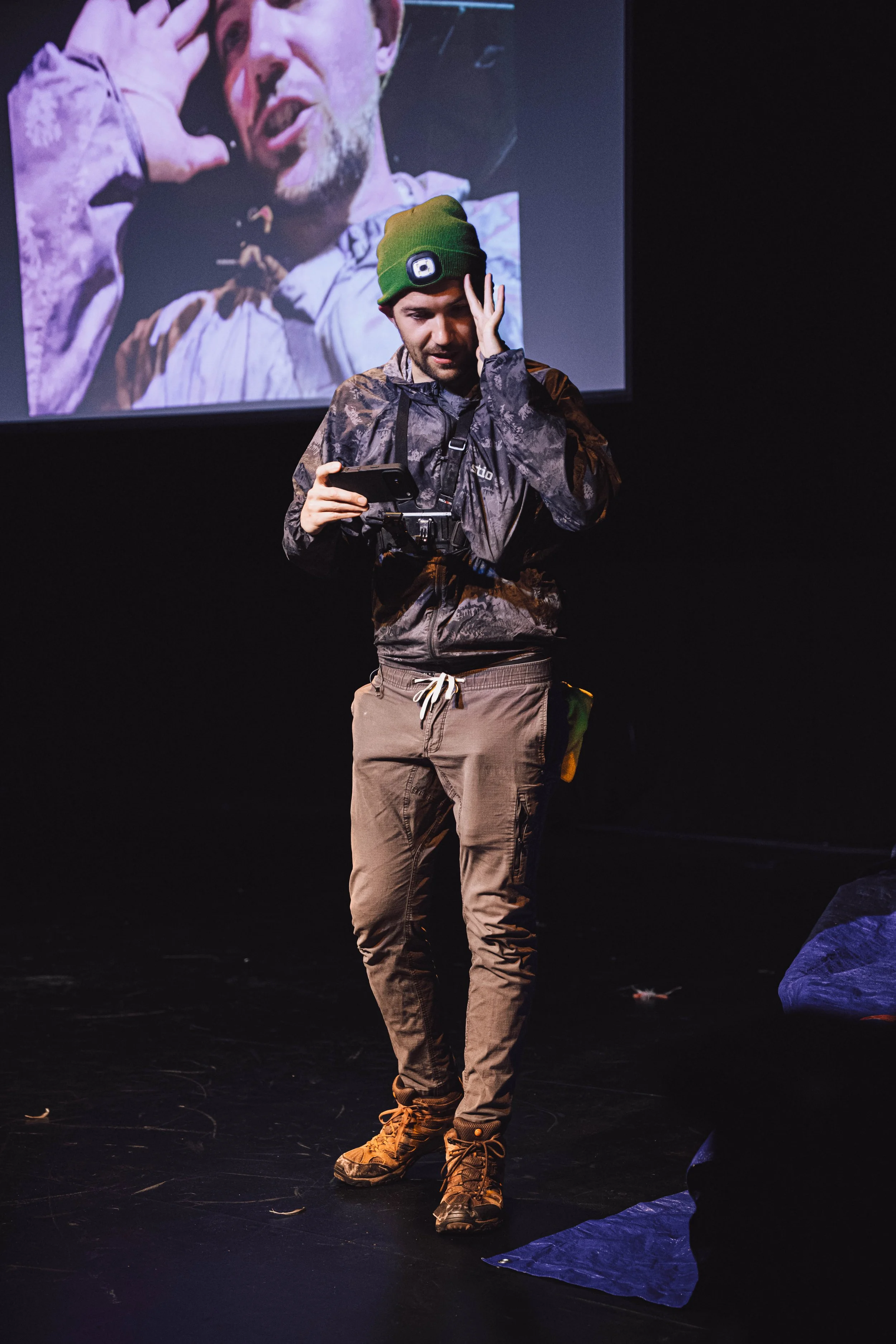 A man in camouflage jacket and beige pants, wearing a green beanie, standing on a stage with a large screen behind him displaying his image. He is holding a phone in one hand and touching his head with the other.