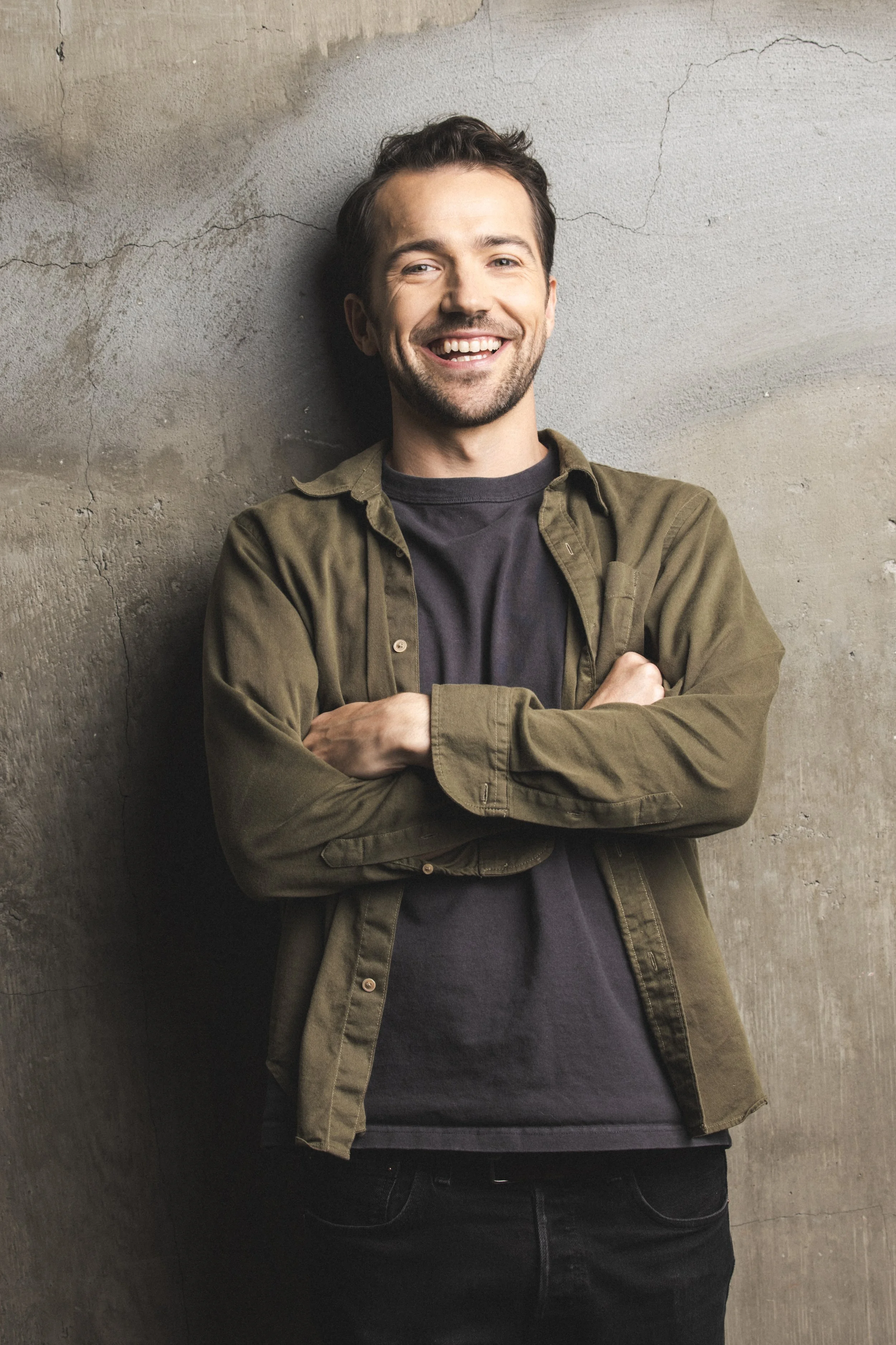 A smiling man with dark hair and a beard, wearing a dark shirt and an olive-green jacket, standing with arms crossed against a textured concrete wall.