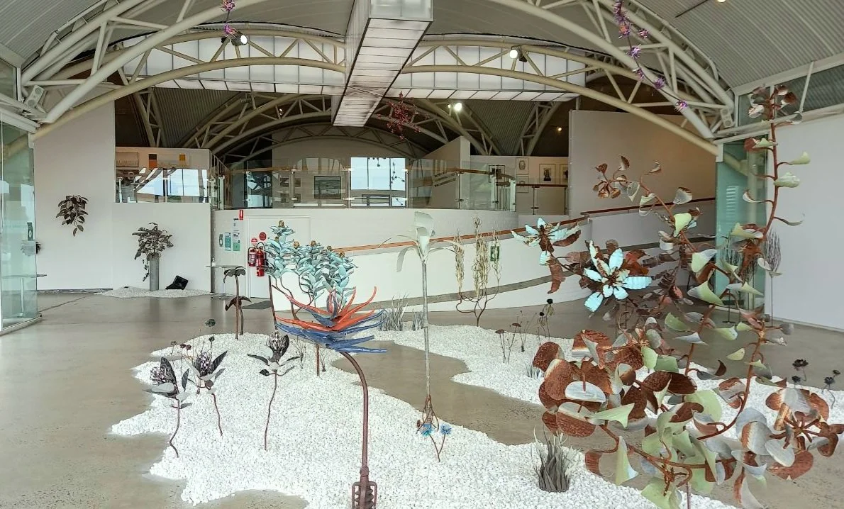 Interior of a modern gallery with art installations of metal plants and flowers in a landscaped area with white gravel, glass walls, and a curved ceiling.