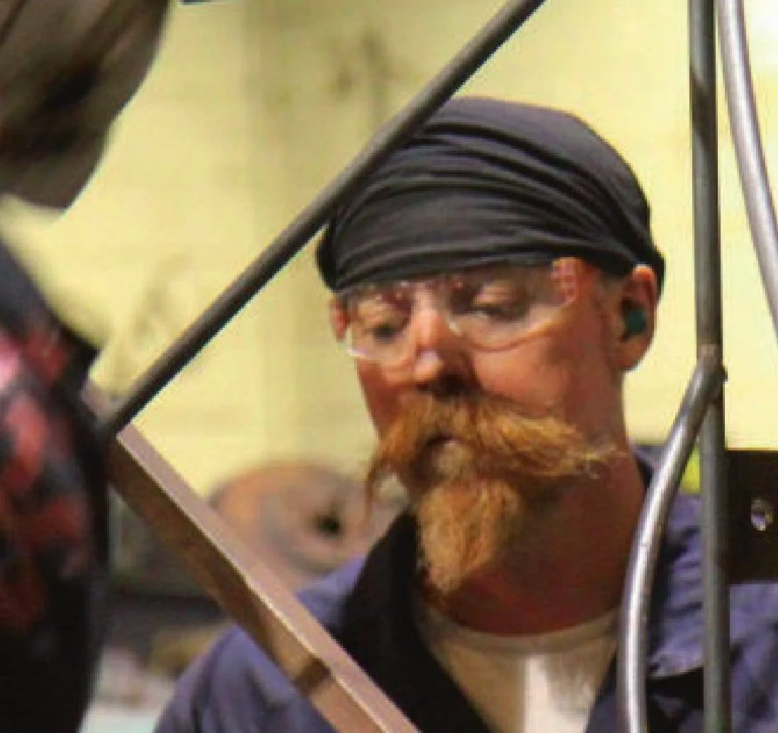Close-up of a man wearing safety glasses, a black head wrap, and a dark jacket, working with metal tools or equipment in an industrial or workshop setting.