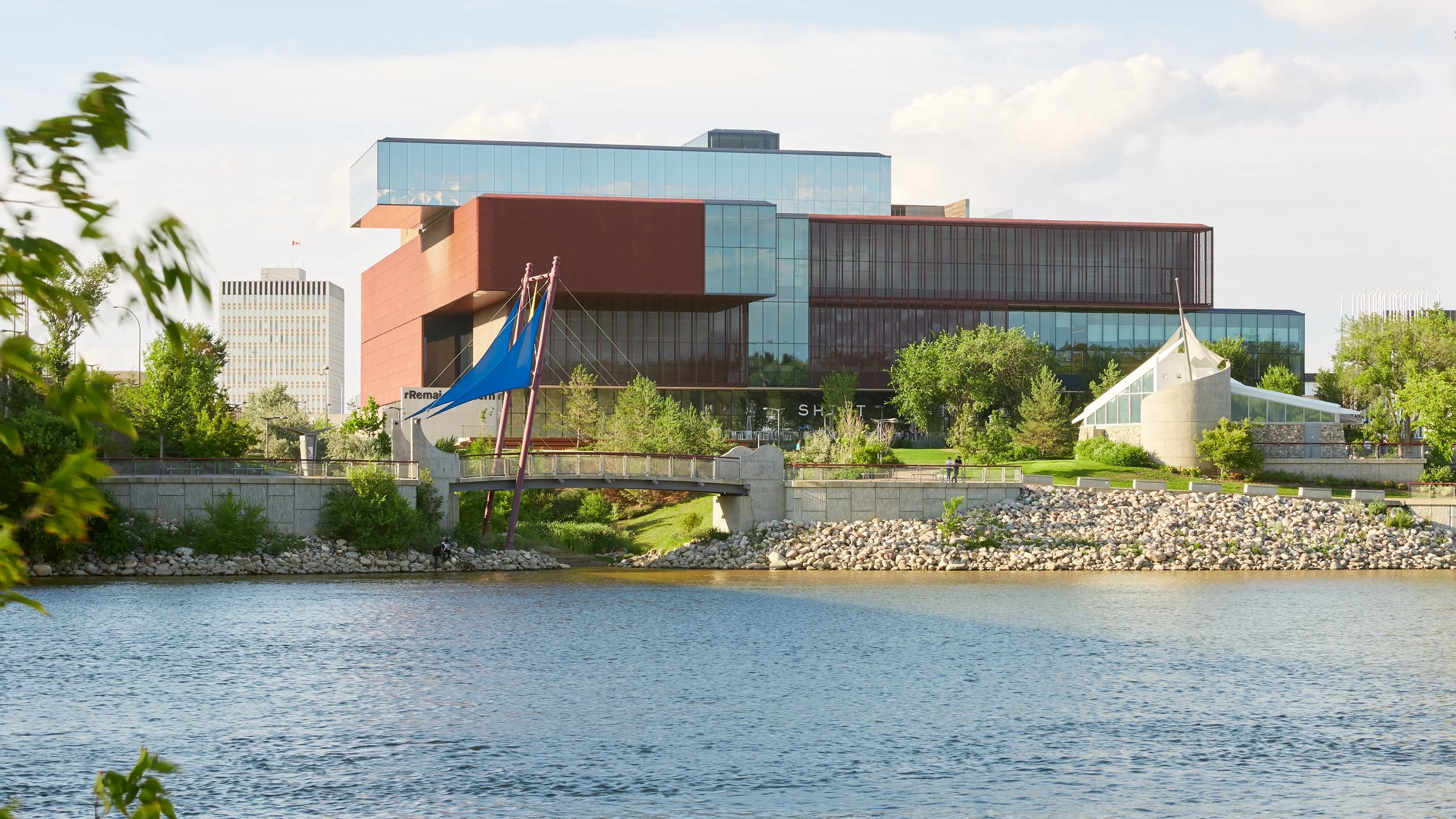 South facade of the Remai Modern Art Gallery in Saskatoon, Saskatchewan. View from across the river. Gallery perched on riverbank above the park. River and walking path in foreground.