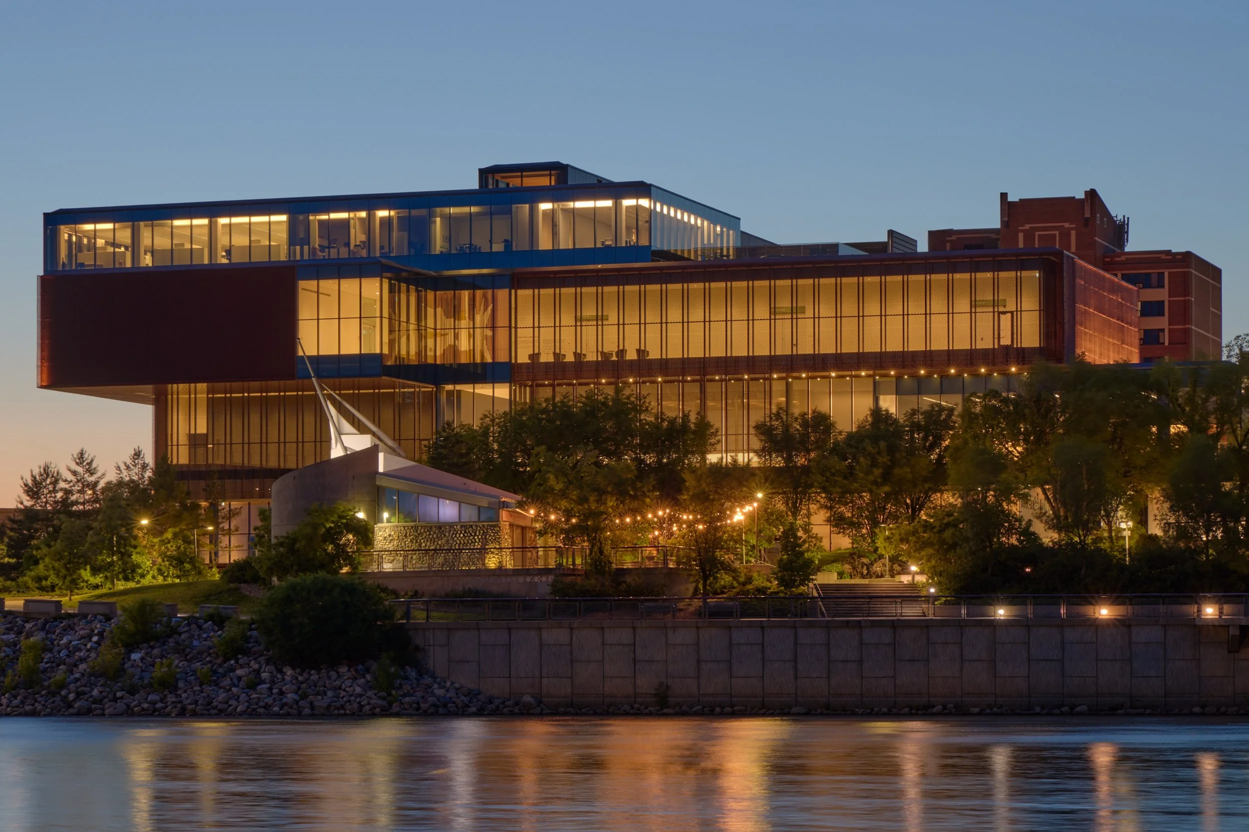 South facade of the Remai Modern Art Gallery in Saskatoon, Saskatchewan. View from across the river. Gallery perched on riverbank above the park. River and walking path in foreground.
