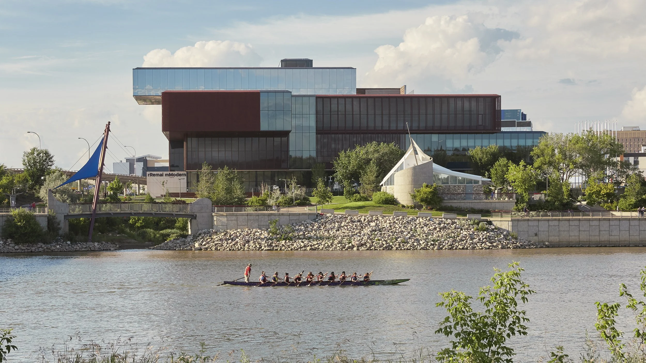 South facade of the Remai Modern Art Gallery in Saskatoon, Saskatchewan. View from across the river. Gallery perched on riverbank above the park, dragonboat team practicing on the river in foreground.