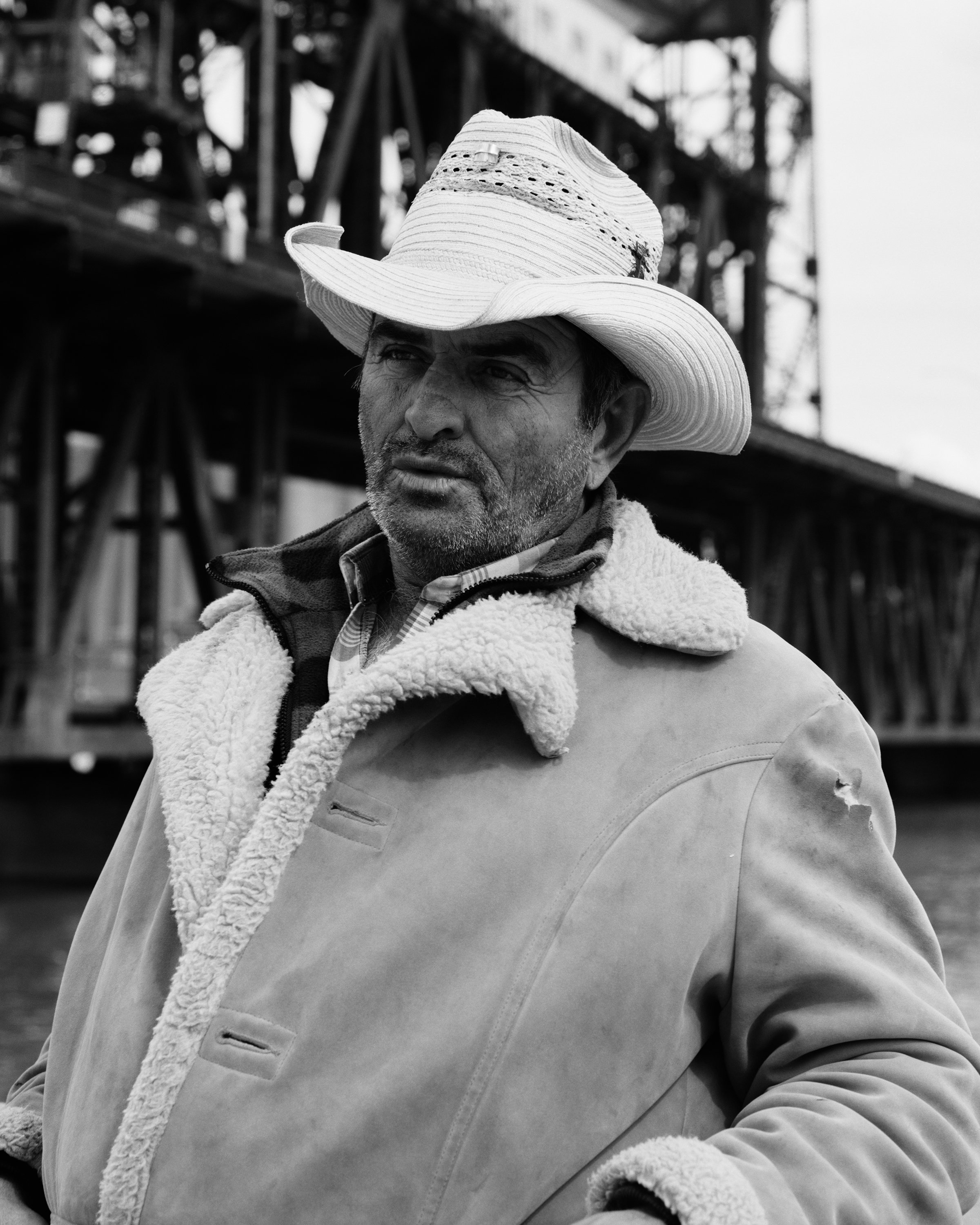Black and white photograph of a man in a sheep and leather jacket and white cowboy hat, leaning against a rail in front of the Steel Bridge in Portland, Oregon.