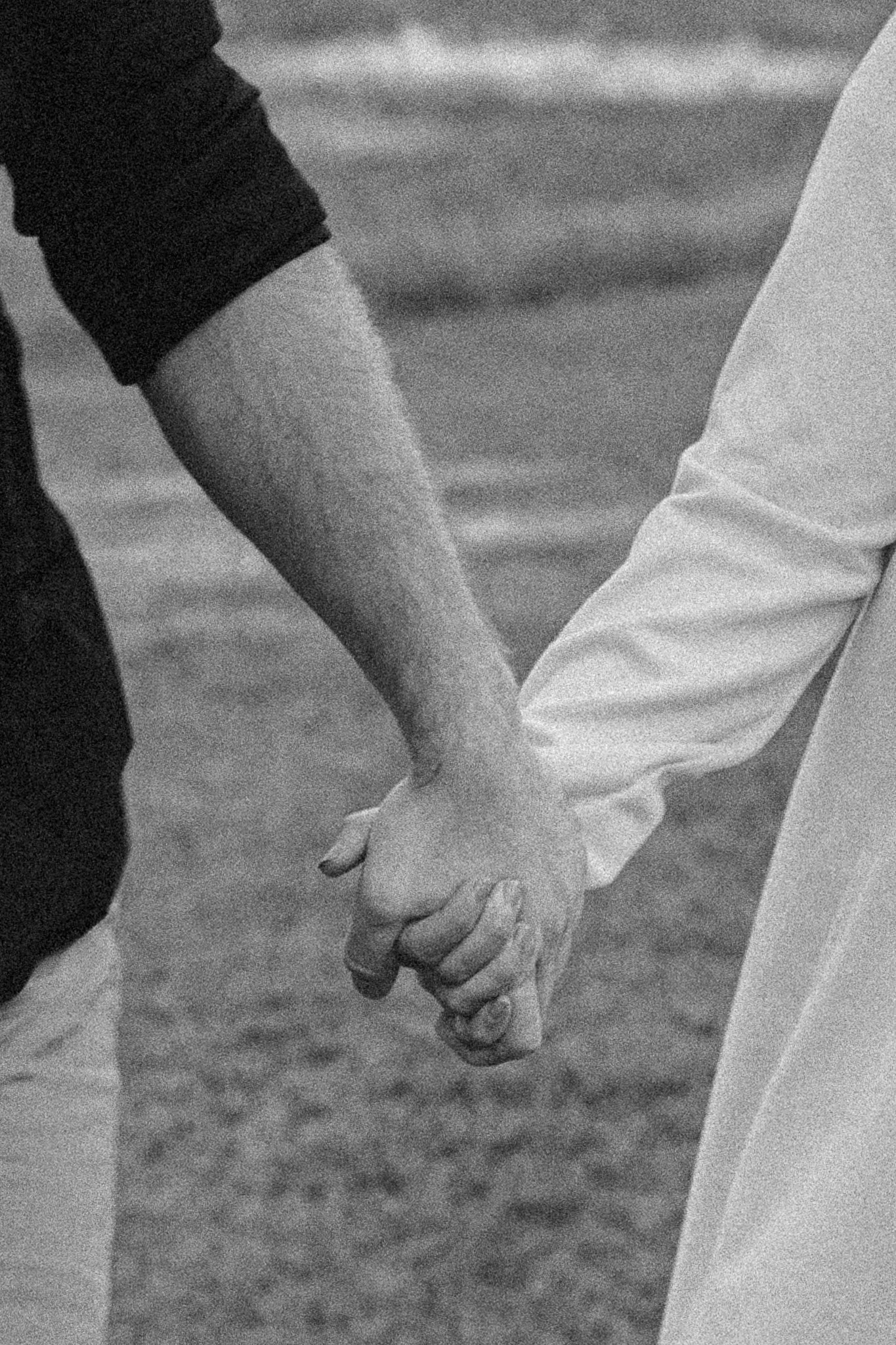 Two people holding hands, one in a dark short-sleeve shirt and the other in a white long-sleeve shirt, against a blurred background.