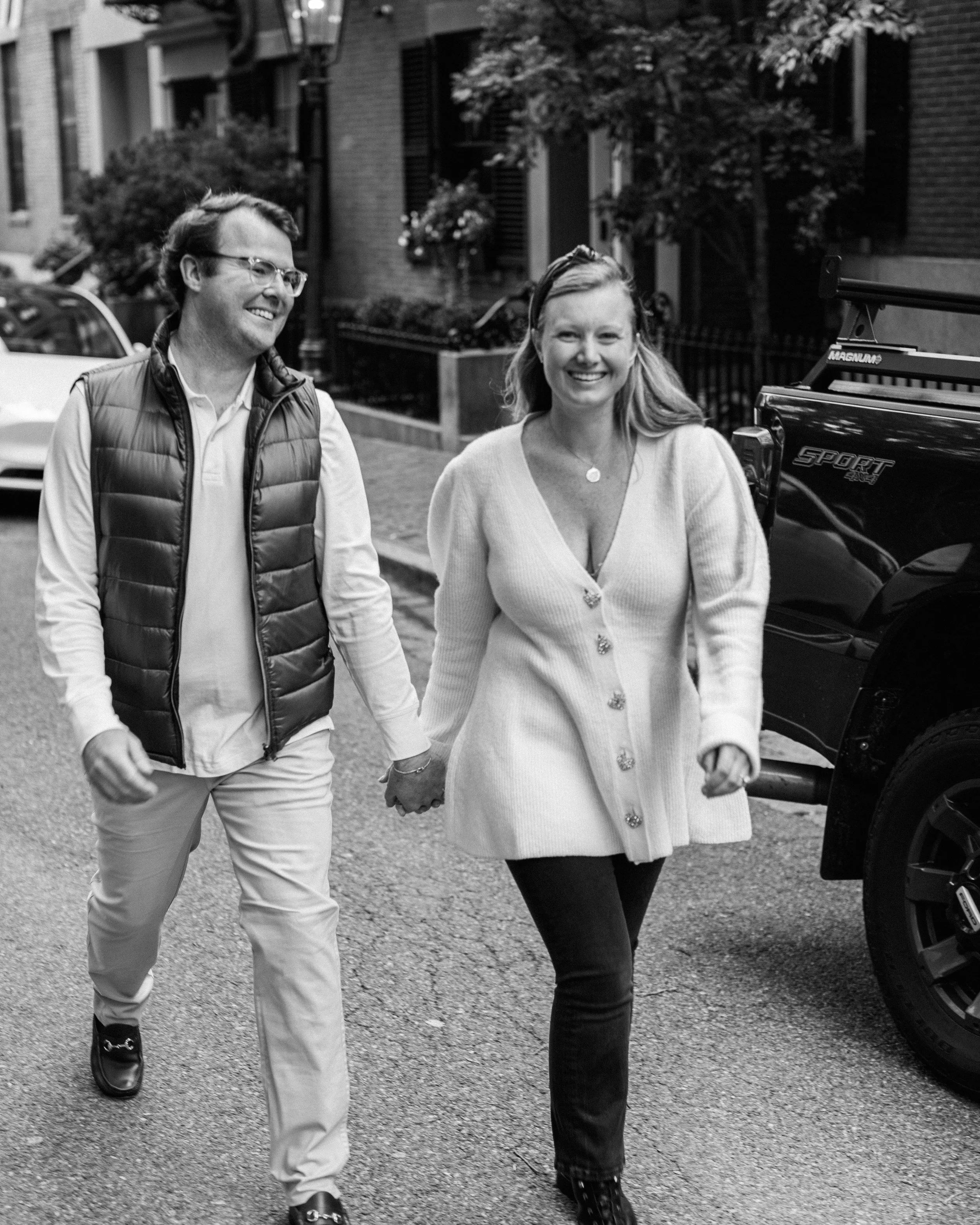 A smiling man and woman holding hands and walking on a city street, with buildings, trees, and parked cars in the background.