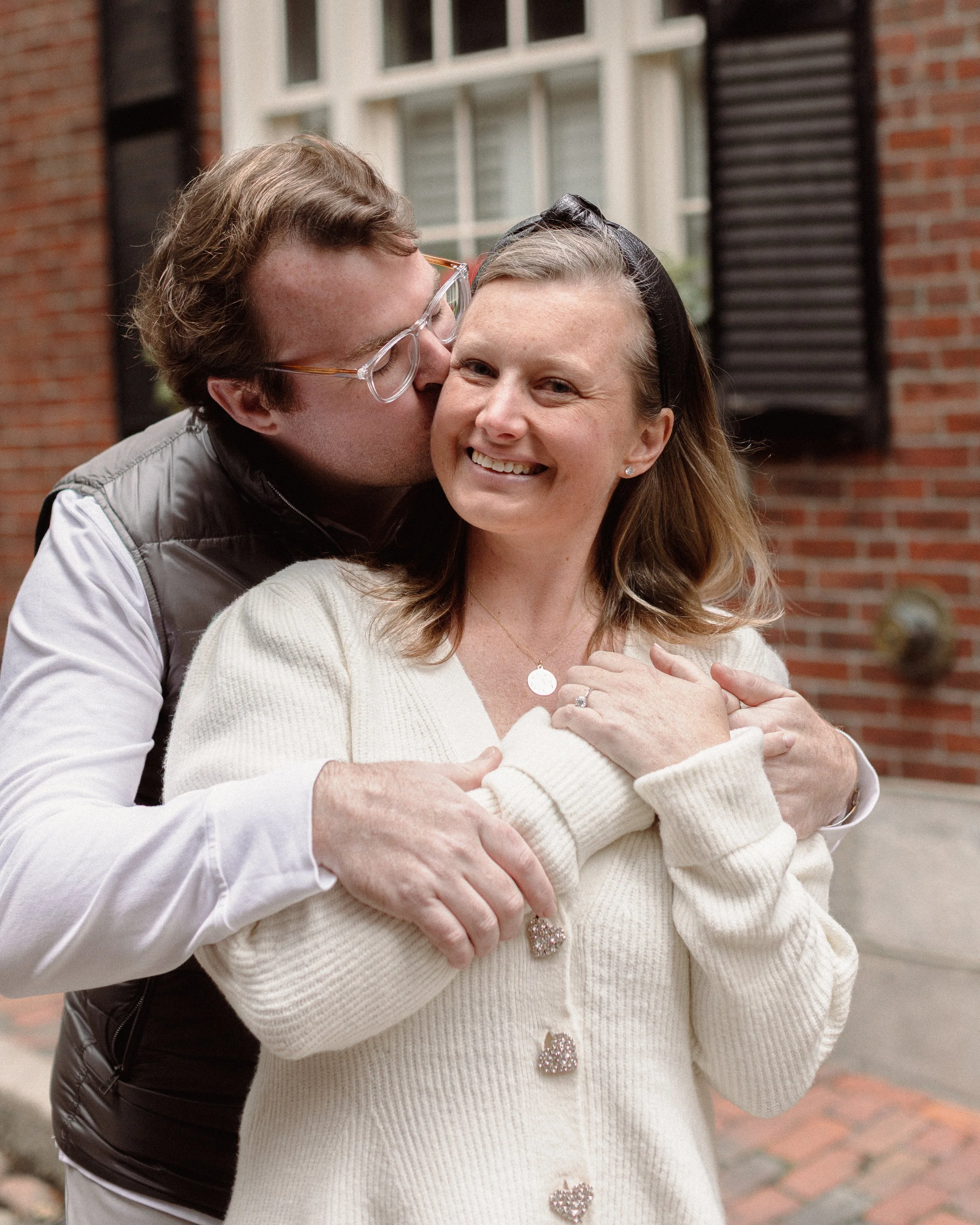 A smiling couple at a celebration. The man has short light brown hair and is wearing a white shirt. The woman has long dark hair, is wearing a white dress with lace details on the shoulders, and has earrings. They are close together, with the woman's arm around the man's shoulders, in a setting with wooden barrels in the background.