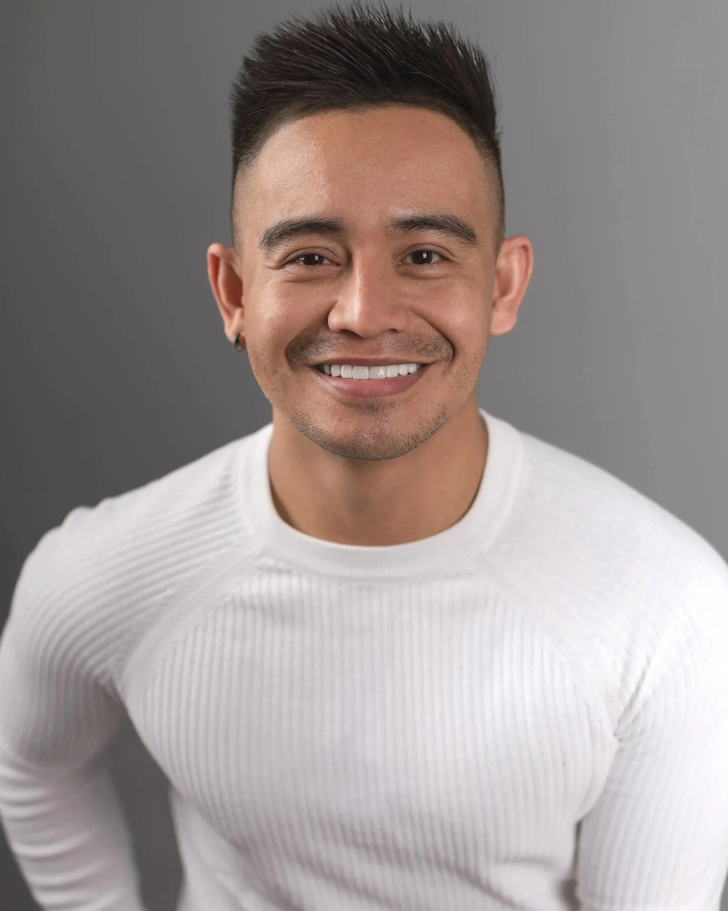 A smiling young man with short dark hair, wearing a white ribbed long-sleeve sweater and dark pants, standing against a plain wall.
