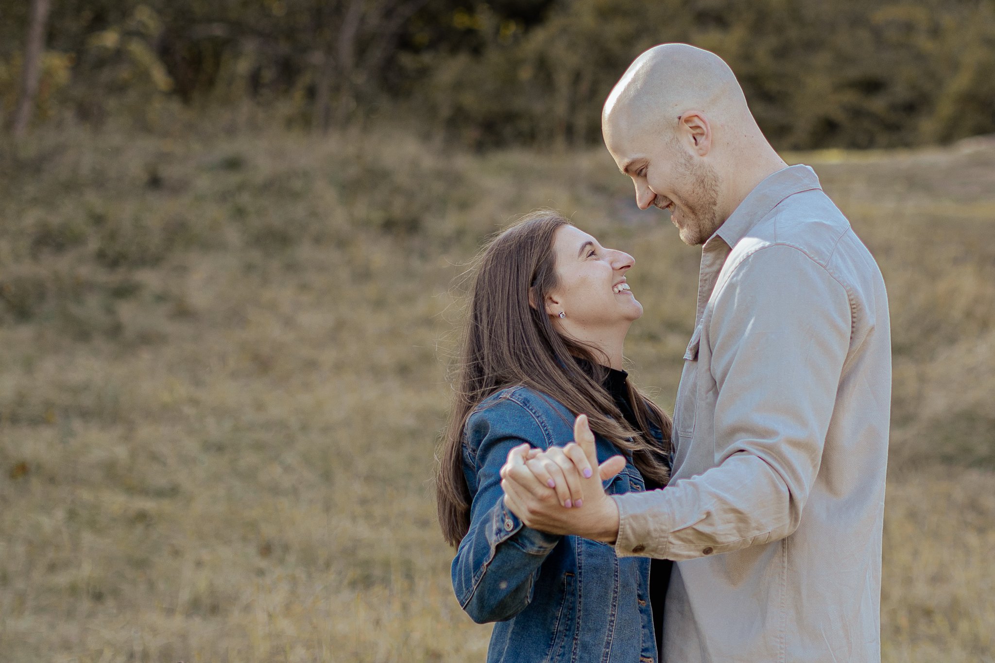 A couple holding hands and looking at each other, smiling, outdoors in a grassy field with trees in the background.