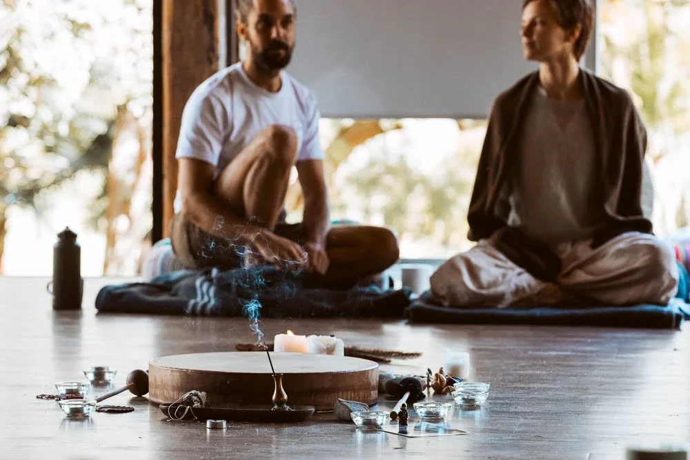 Two individuals sitting cross-legged on a wooden floor in a room with large windows, with a burning candle and various small objects on the floor in front of them, possibly practicing meditation or spiritual rituals.