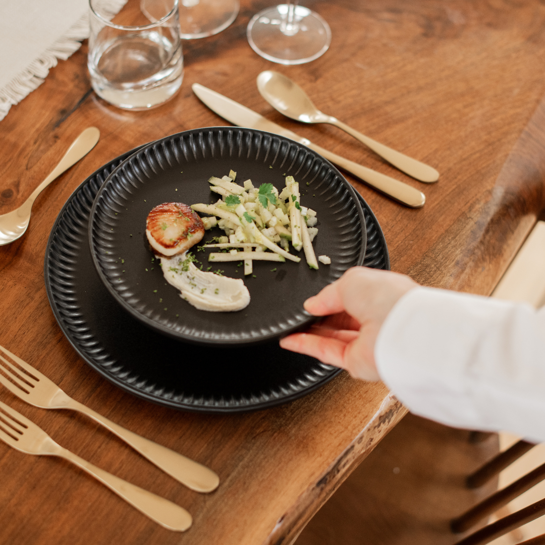 A person holds a black plate with a scallop, a dollop of white sauce, and a side of sliced fennel garnished with herbs, on a wooden dining table with gold utensils, glasses, and a water glass.