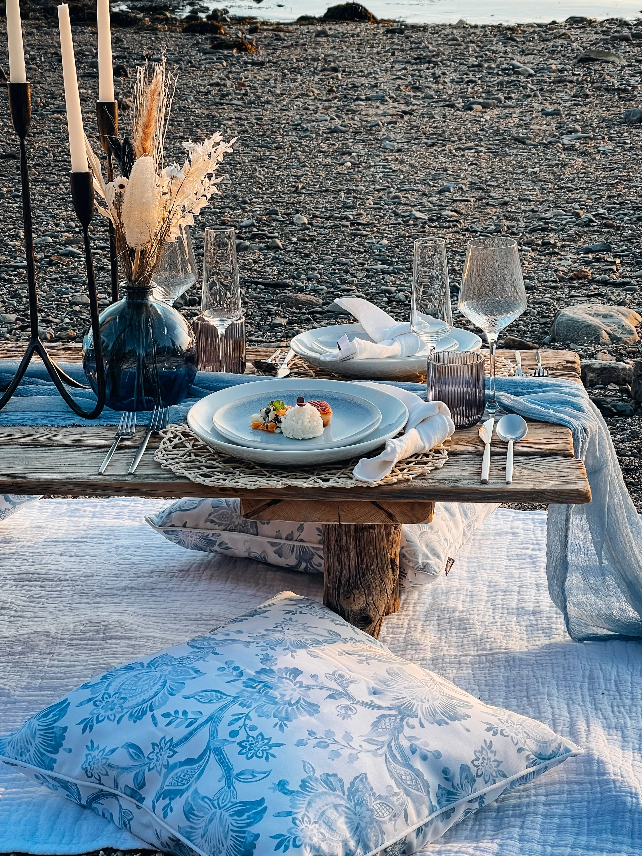 A beachside outdoor dining setup with a low wooden table, white plate with food, glassware, and decorative candle holder. Cushions and blankets are on the rocky beach, creating a cozy, elegant scene at sunset.